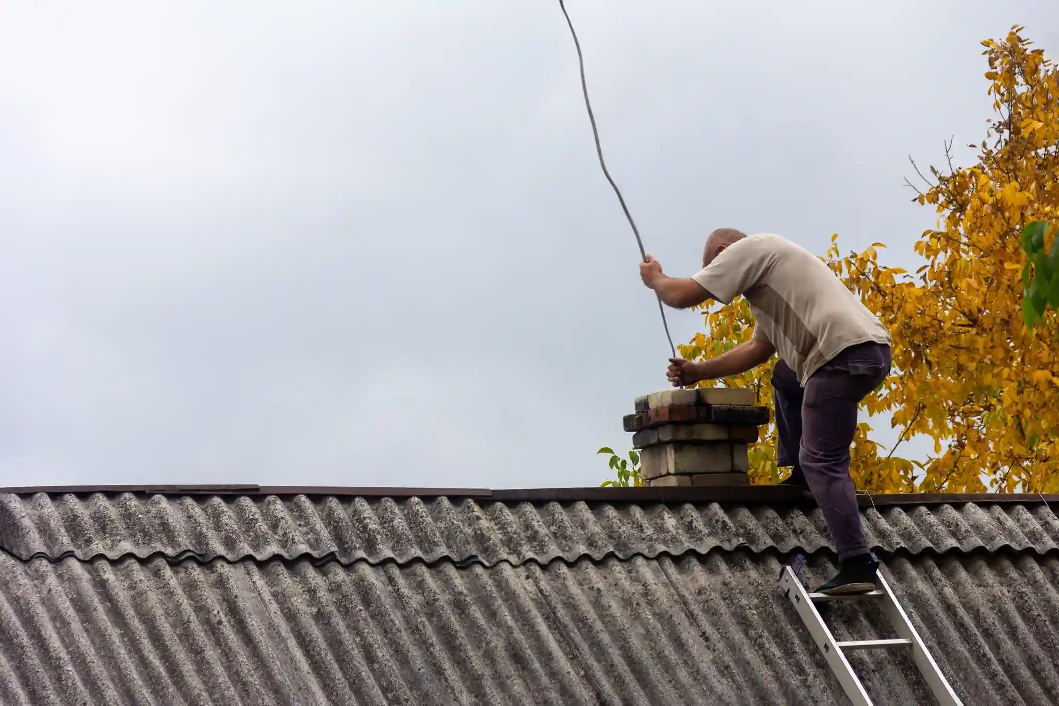 Technician standing on a ladder on an old roof, feeding a chimney cleaning rod into the brick chimney.