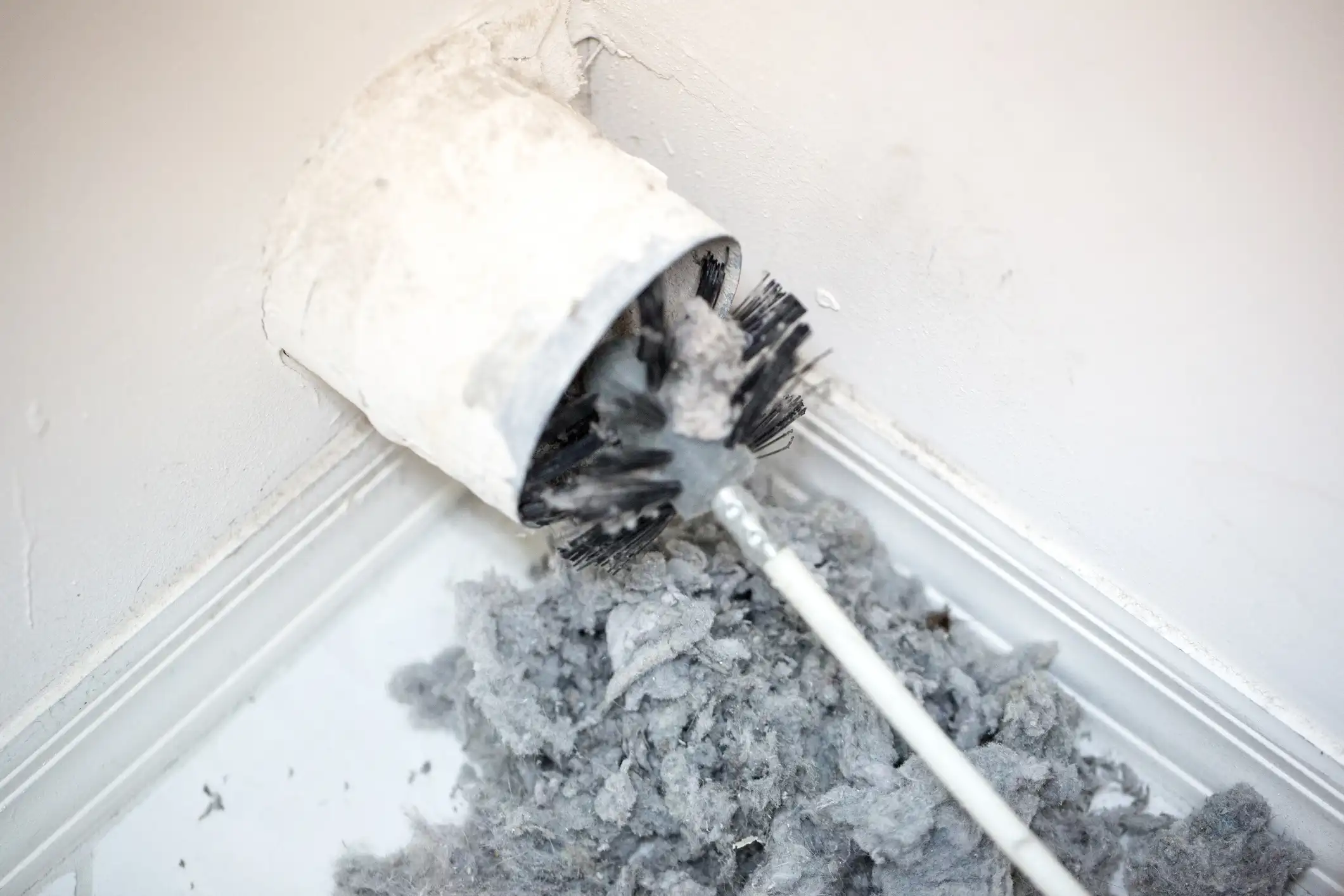  A close-up, high-angle view shows a long, white-handled brush with black bristles being inserted into a white exhaust vent pipe coming out of a wall corner. The brush is covered in thick, light gray lint. A large pile of the same gray lint lies below the vent, indicating a recent or ongoing dryer vent cleaning process. The surrounding wall and trim are a rough, light color.