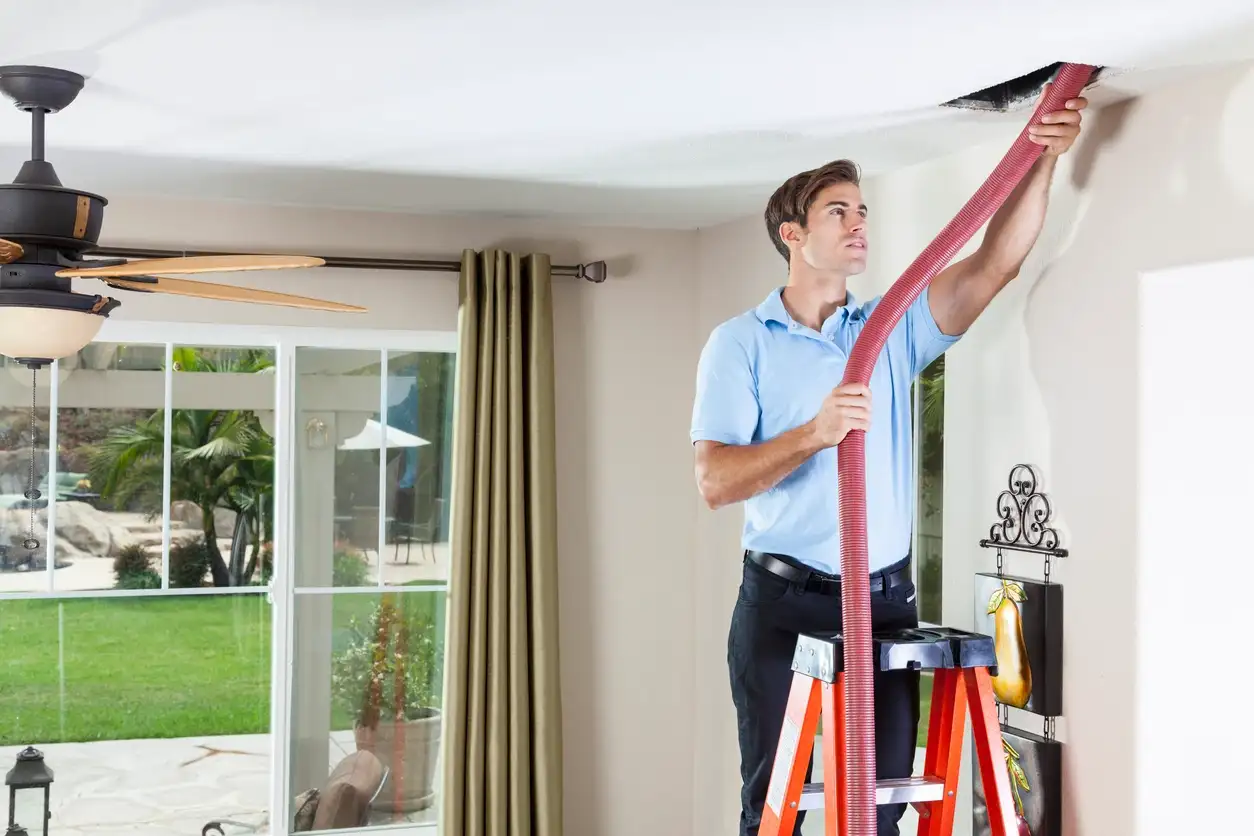 A male technician on a ladder in a living room uses a large red vacuum hose to clean a rectangular air vent in a ceiling.