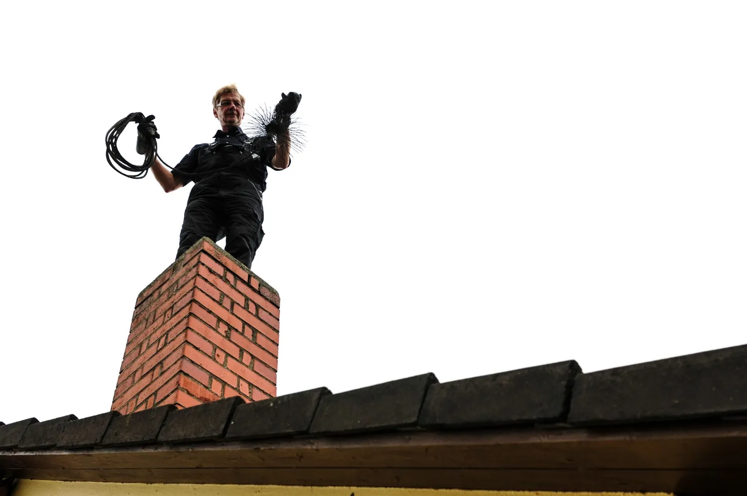 A chimney sweep standing atop a square, red brick chimney stack. The worker, who has light-colored hair, is dressed in a dark uniform and gloves. In one hand, they hold a coil of black rope or cable, and in the other, a large, bristled chimney brush connected to a rod or chain, ready for cleaning the flue. The roof below consists of dark, overlapping tiles. The figure and chimney are sharply silhouetted against an overwhelmingly bright white sky, creating a high-contrast and elevated perspective.