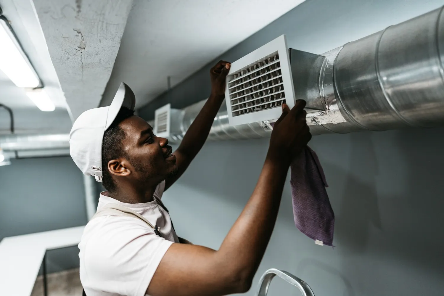 A side view shows a smiling HVAC technician wearing a white baseball cap and overalls, standing on a ladder. He is holding a square white air vent cover with both hands, removing it from or installing it onto a large, cylindrical metal air duct on a gray wall. He holds a purple cleaning cloth in his right hand.