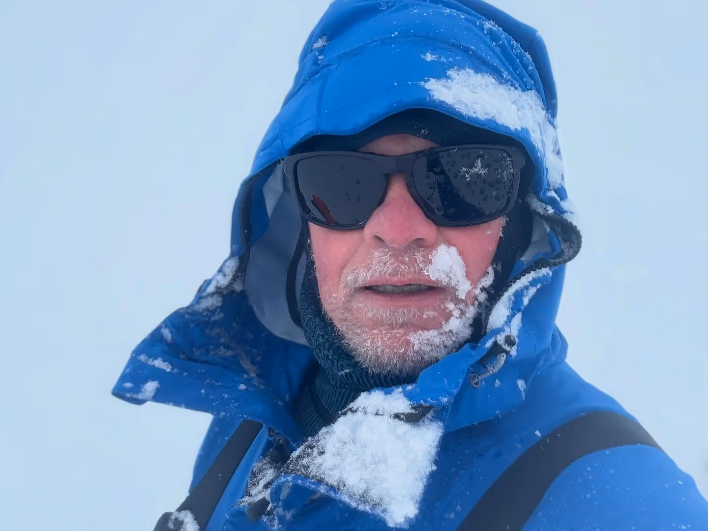 A man wearing a blue coat and sunglasses with snow on his face.