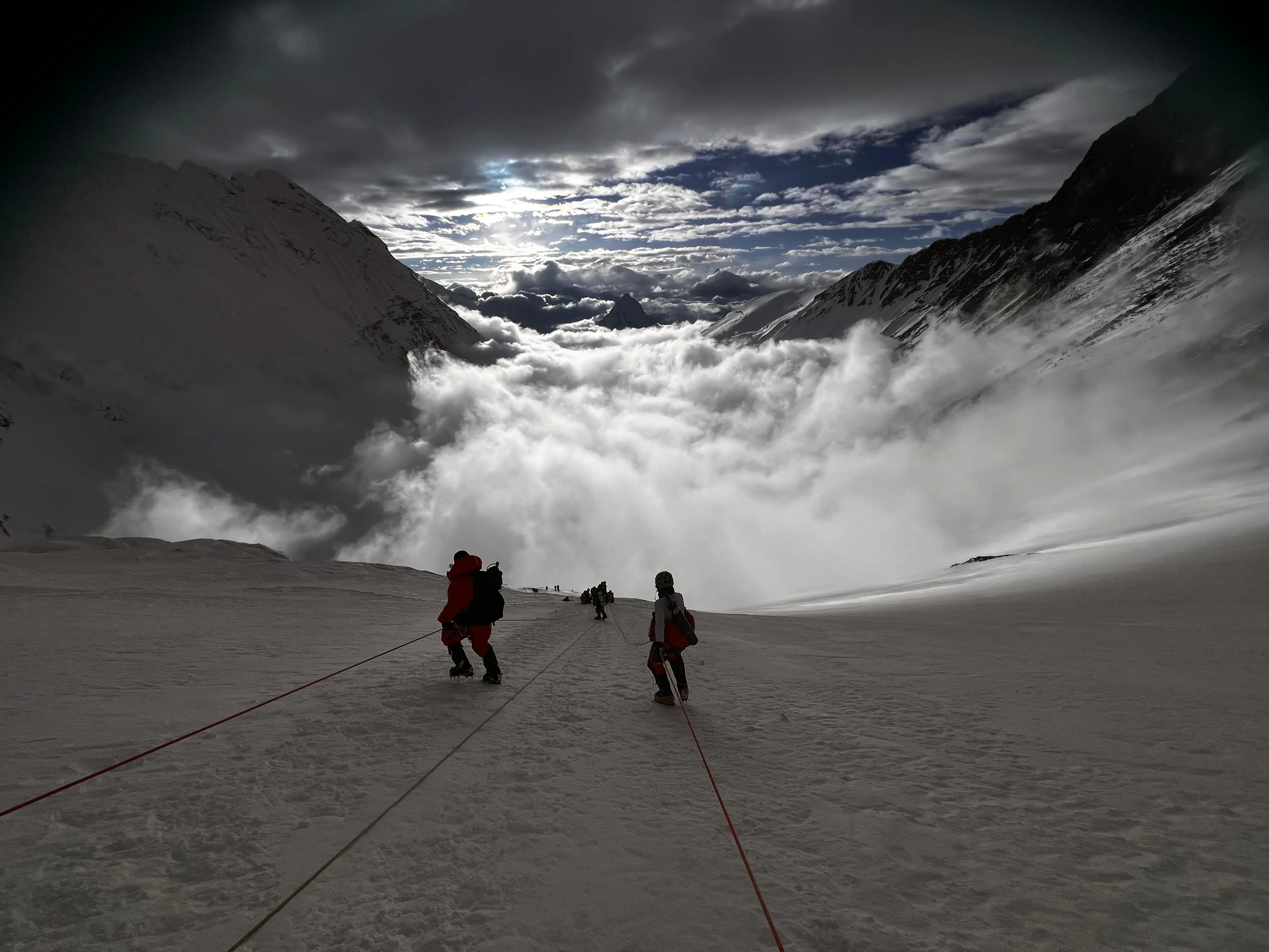 Two roped climbers descending a broad snow slope high on the mountain, with a sea of cloud filling the valley and peaks breaking through in the distance