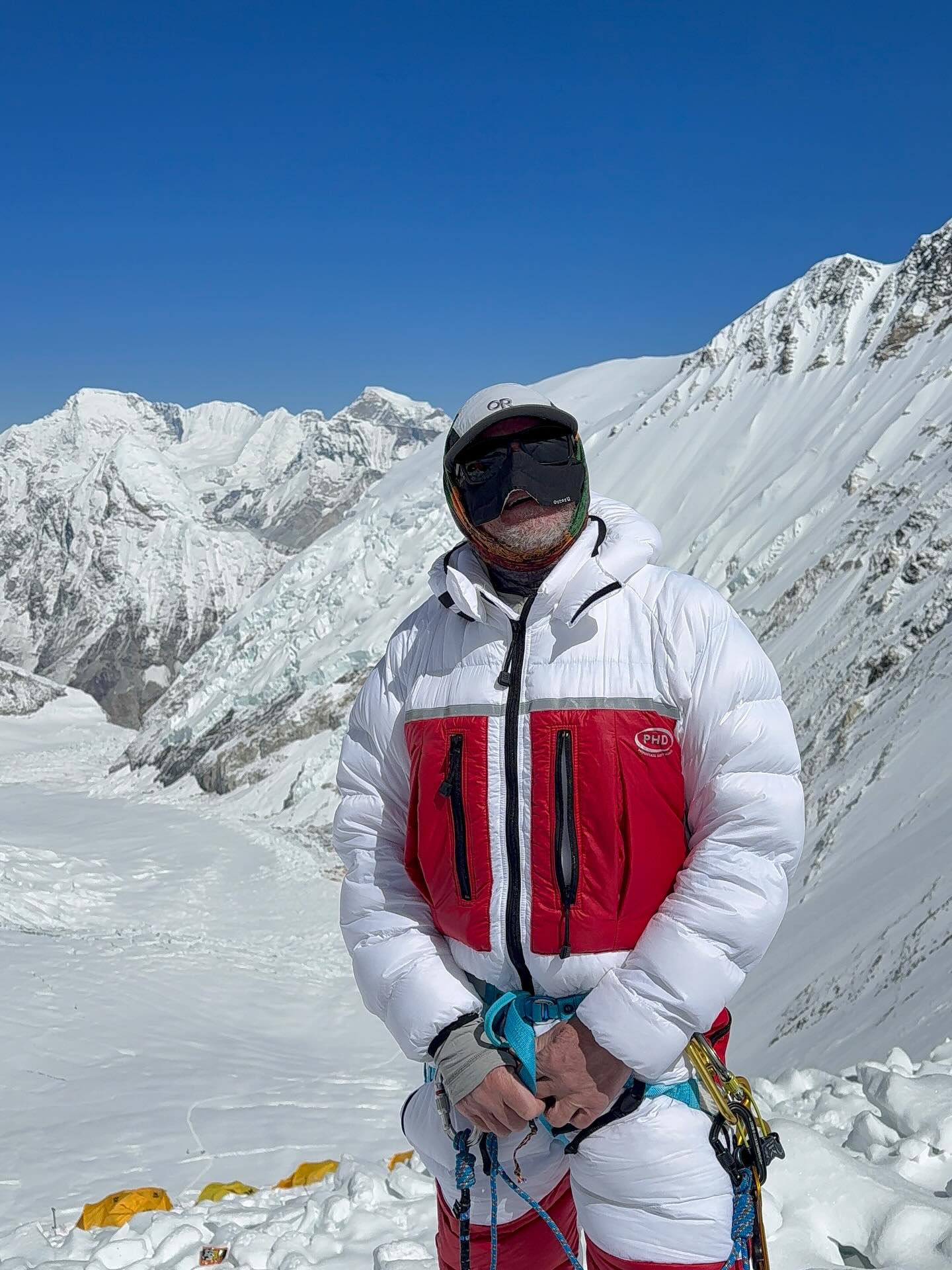 A climber in a red and white down suit and goggles on a snow slope below high peaks