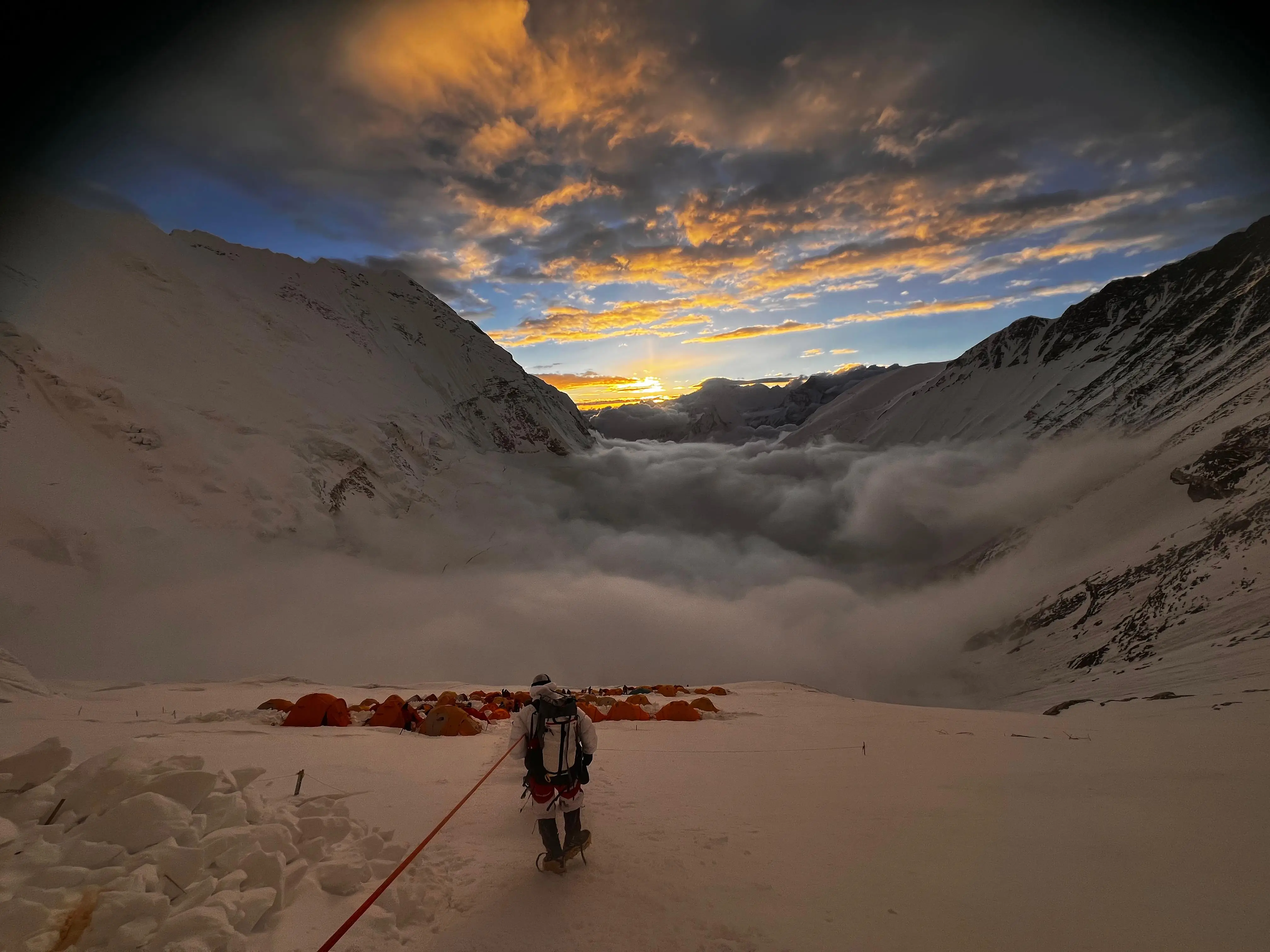 A climber roped up, walking towards a cluster of orange tents at sunset on Everest