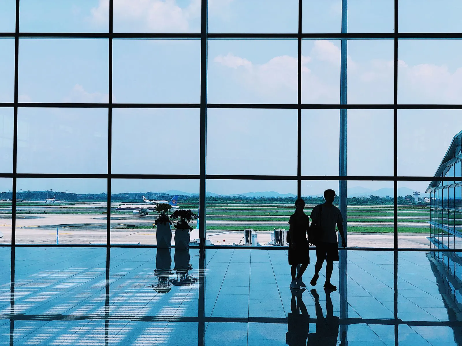Deux personnes marchent dans un terminal d'aéroport avec avions visibles à travers la grande baie vitrée.