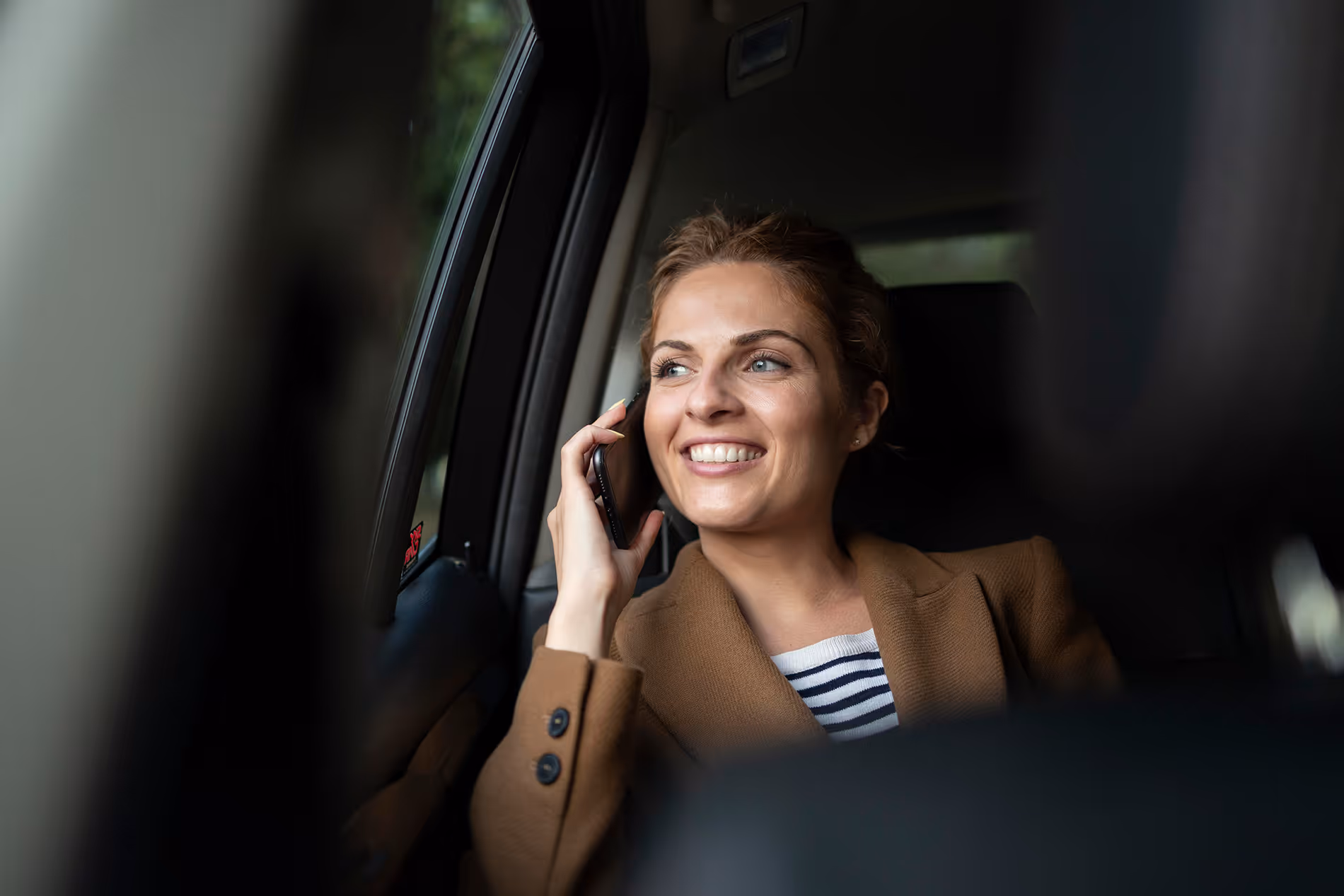 Femme souriante tenant un téléphone assise à l'arrière d'une voiture regardant par la fenêtre.