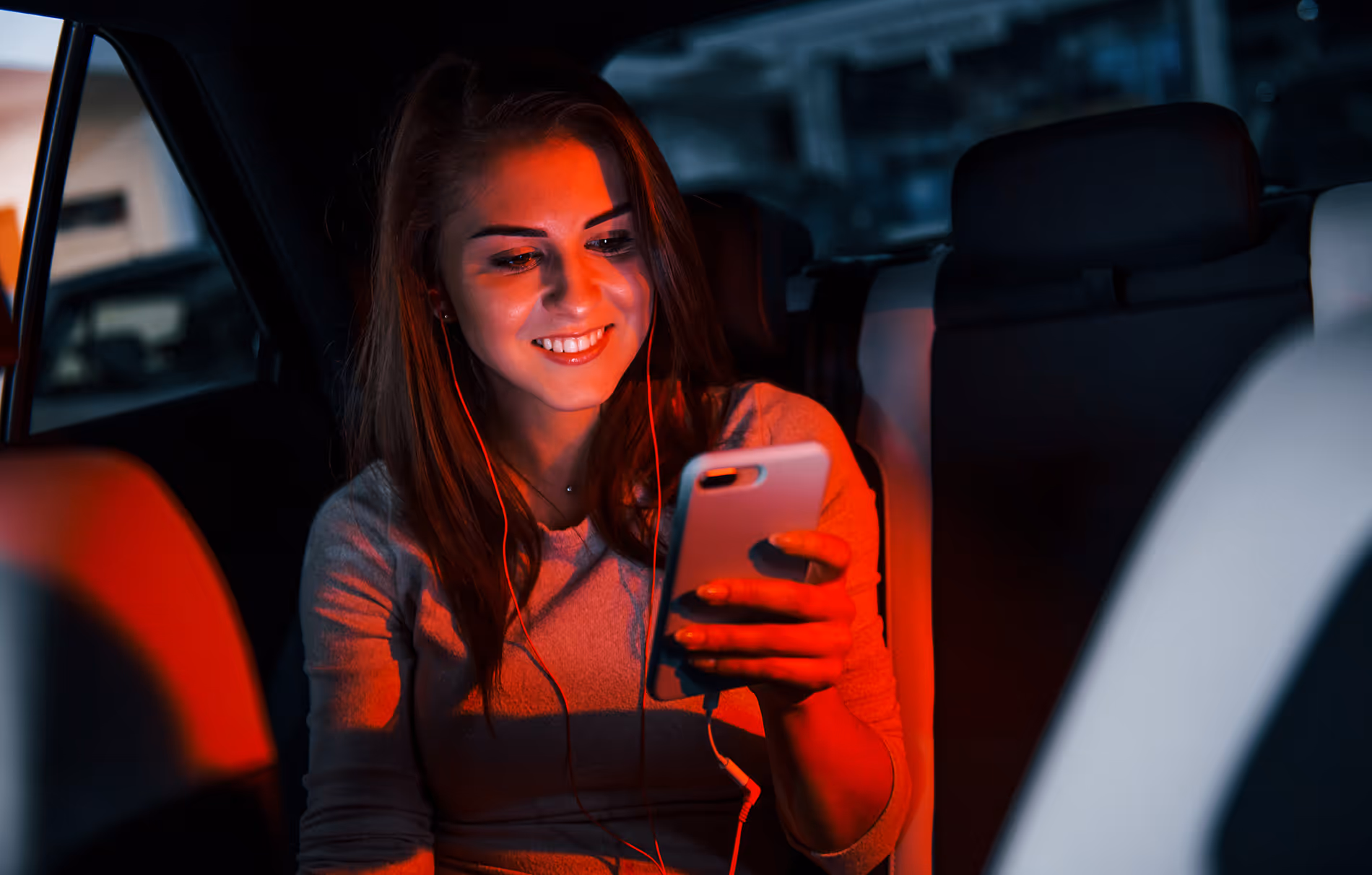 Jeune femme souriante dans une voiture regardant son smartphone avec des écouteurs rouges.