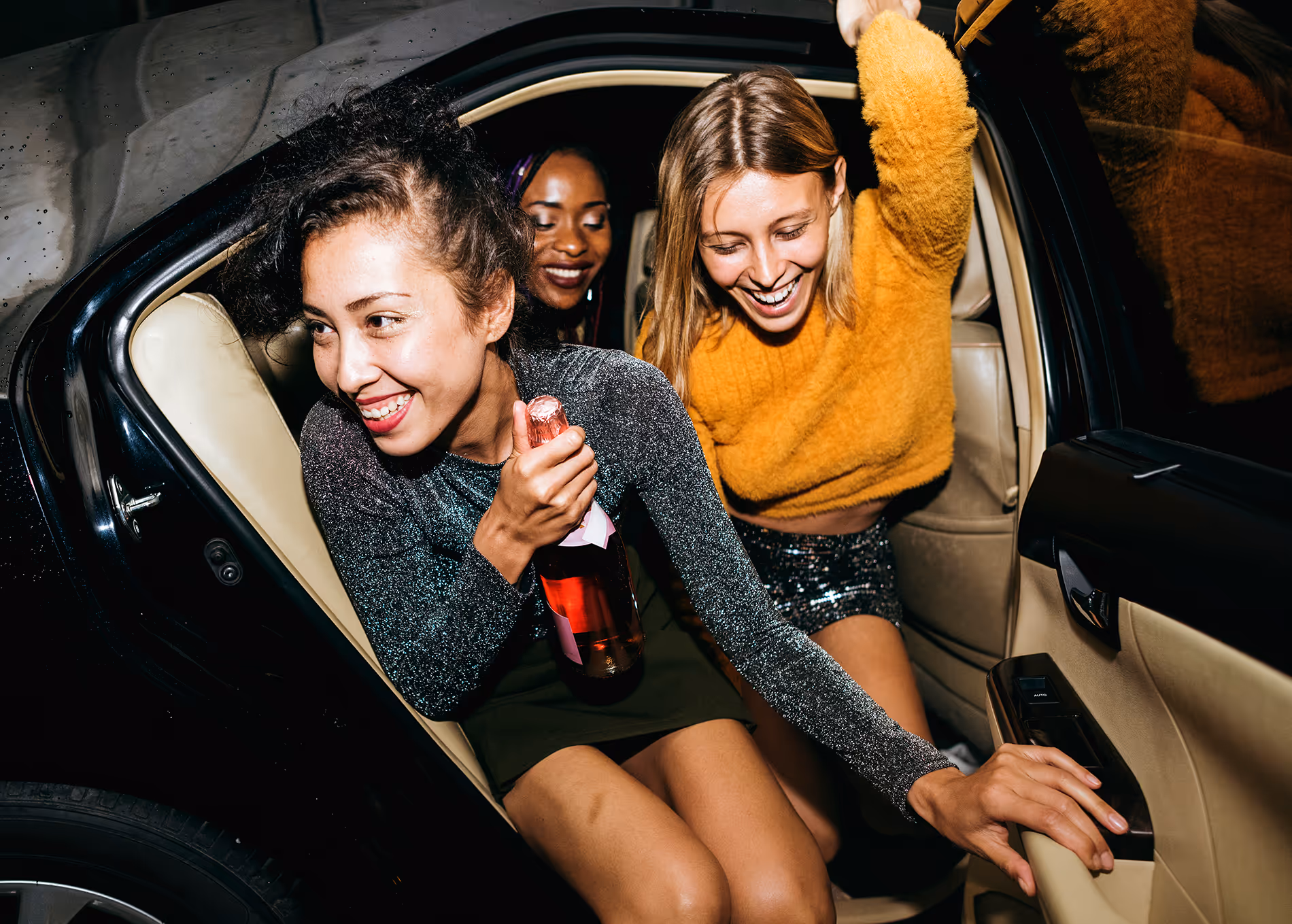 Trois femmes souriantes dans une voiture, l'une tenant une bouteille de vin mousseux.
