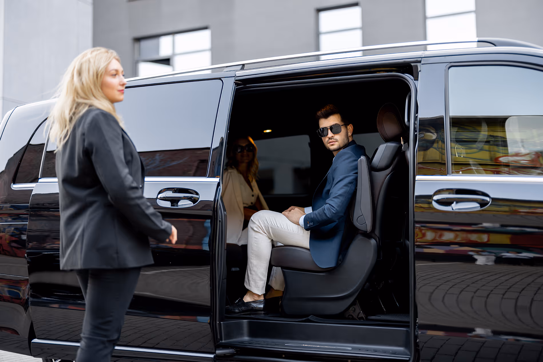 Un homme élégant en lunettes de soleil assis dans une voiture noire avec une femme debout à côté.