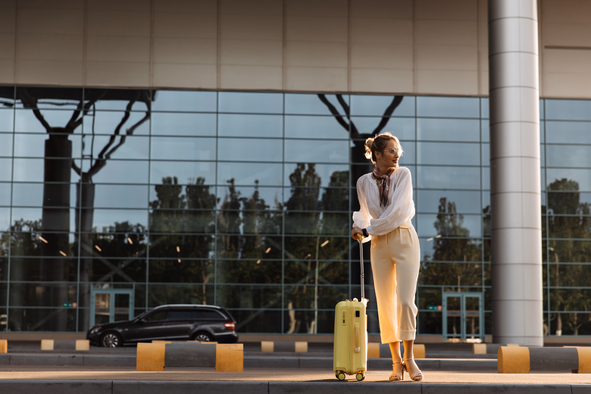 Femme élégante avec valise jaune debout devant une façade vitrée réfléchissante.