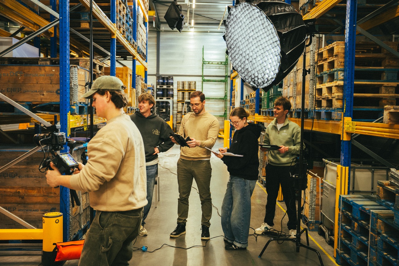 Vijf mensen werken met camera's en tablets in een magazijn met stellingen en pallets.