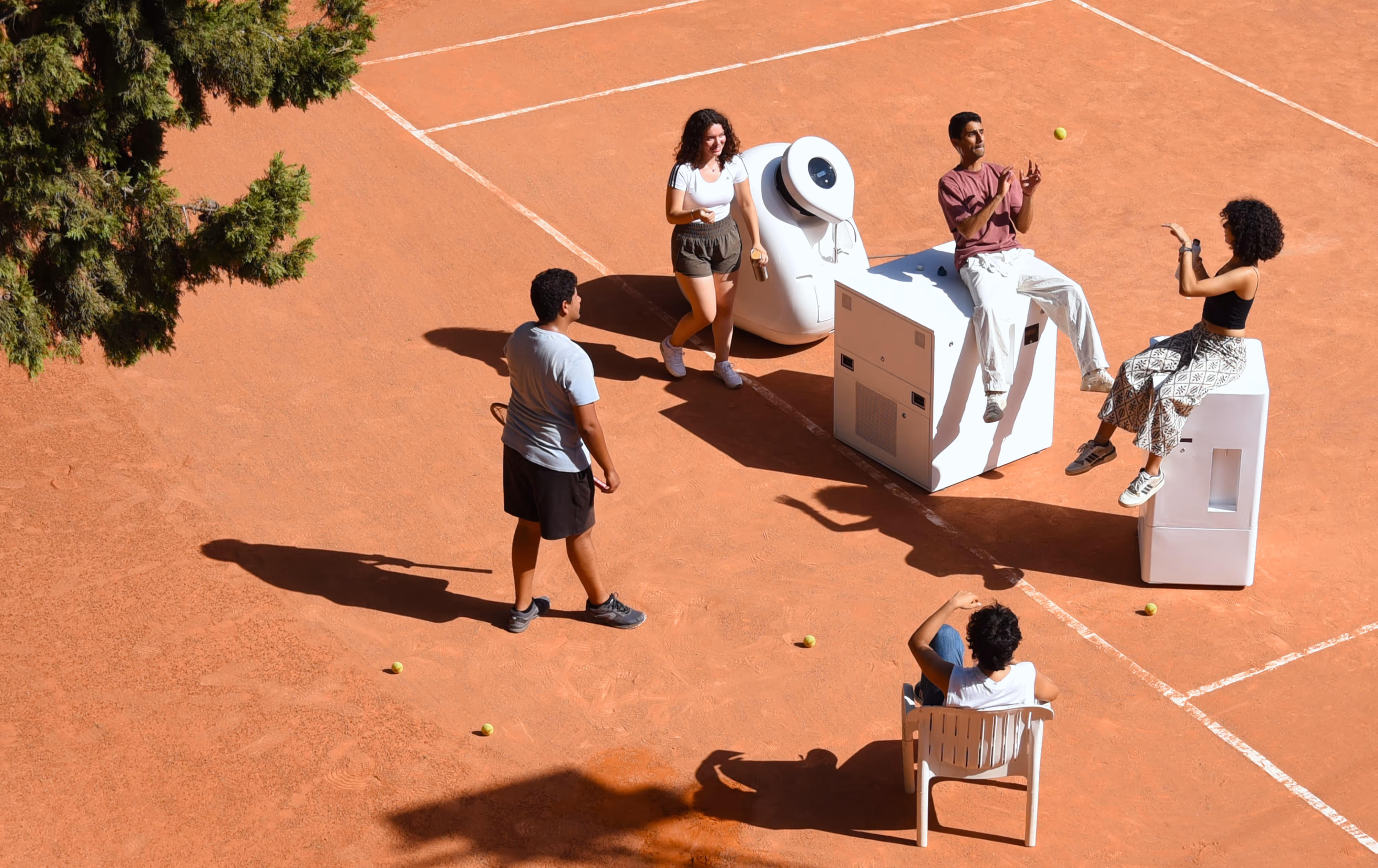 A group of friends standing around a tennis court, next to each model of Kumulus air water generators - namely the Amphore, Boks, and Water Cooler.