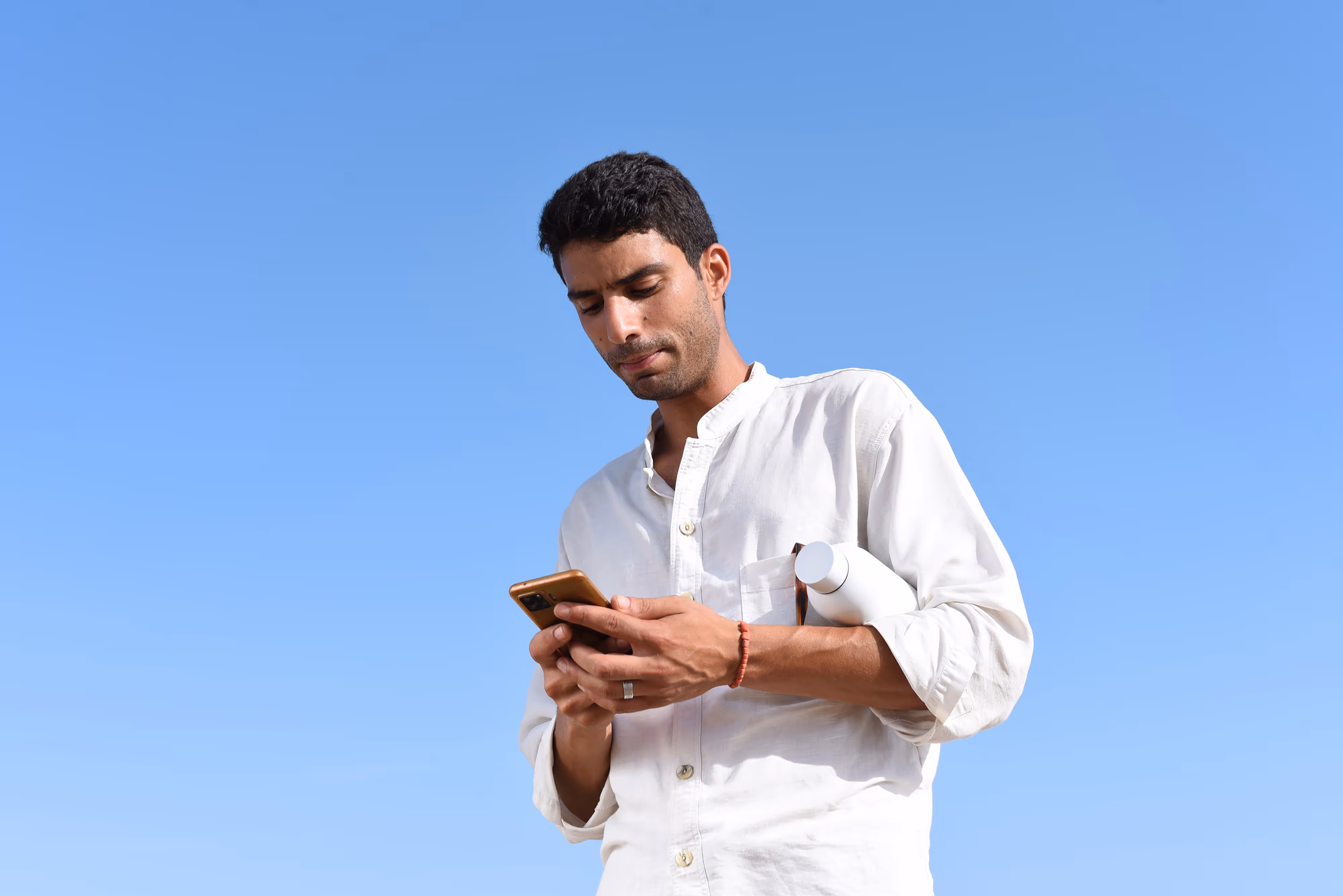 A man checking his phone, while holding a water bottle, against the backdrop of a clear blue sky.