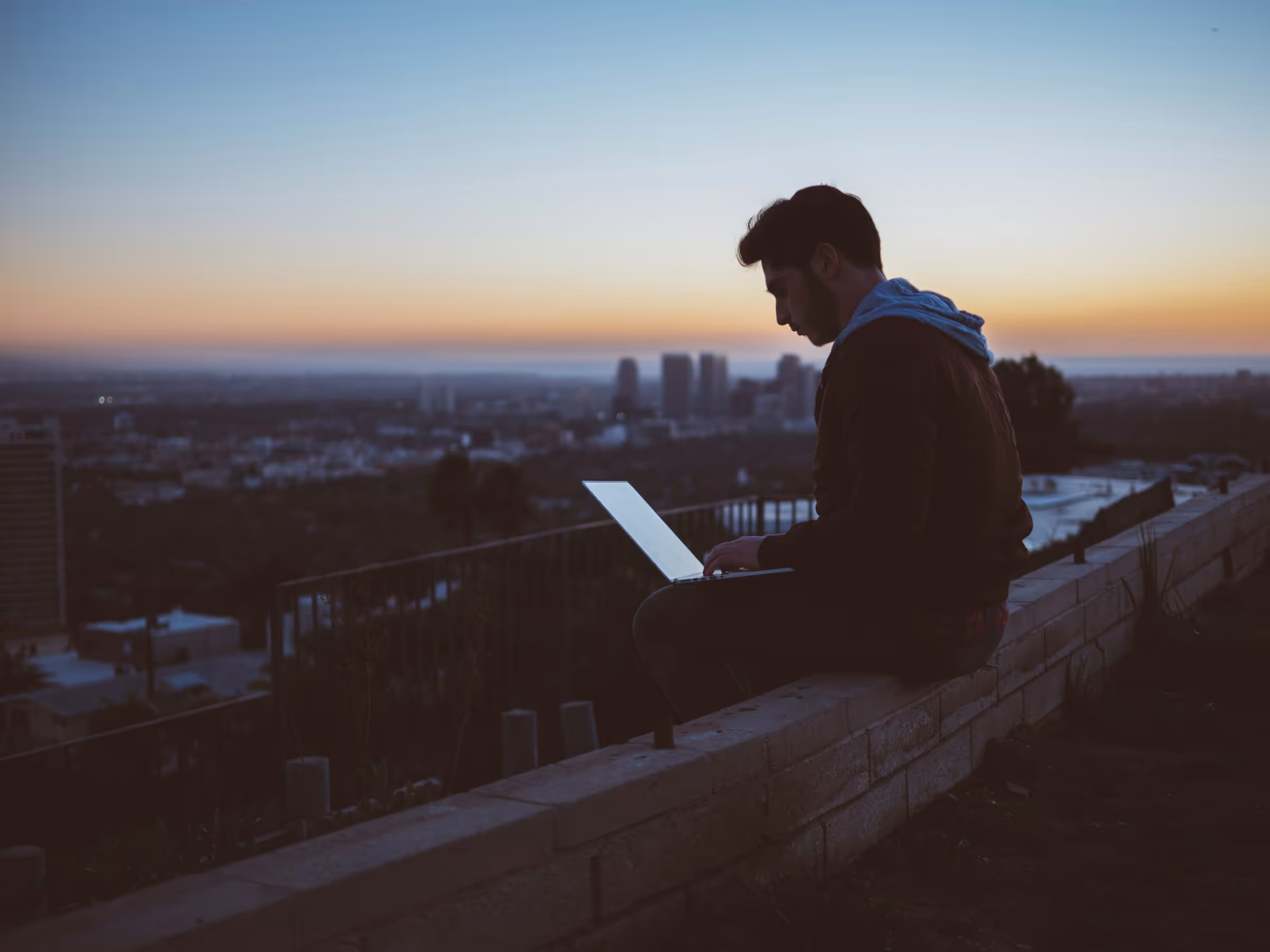 A man sits on the ledge of a building as he's working on his laptop. The background shows a beautiful sunset.