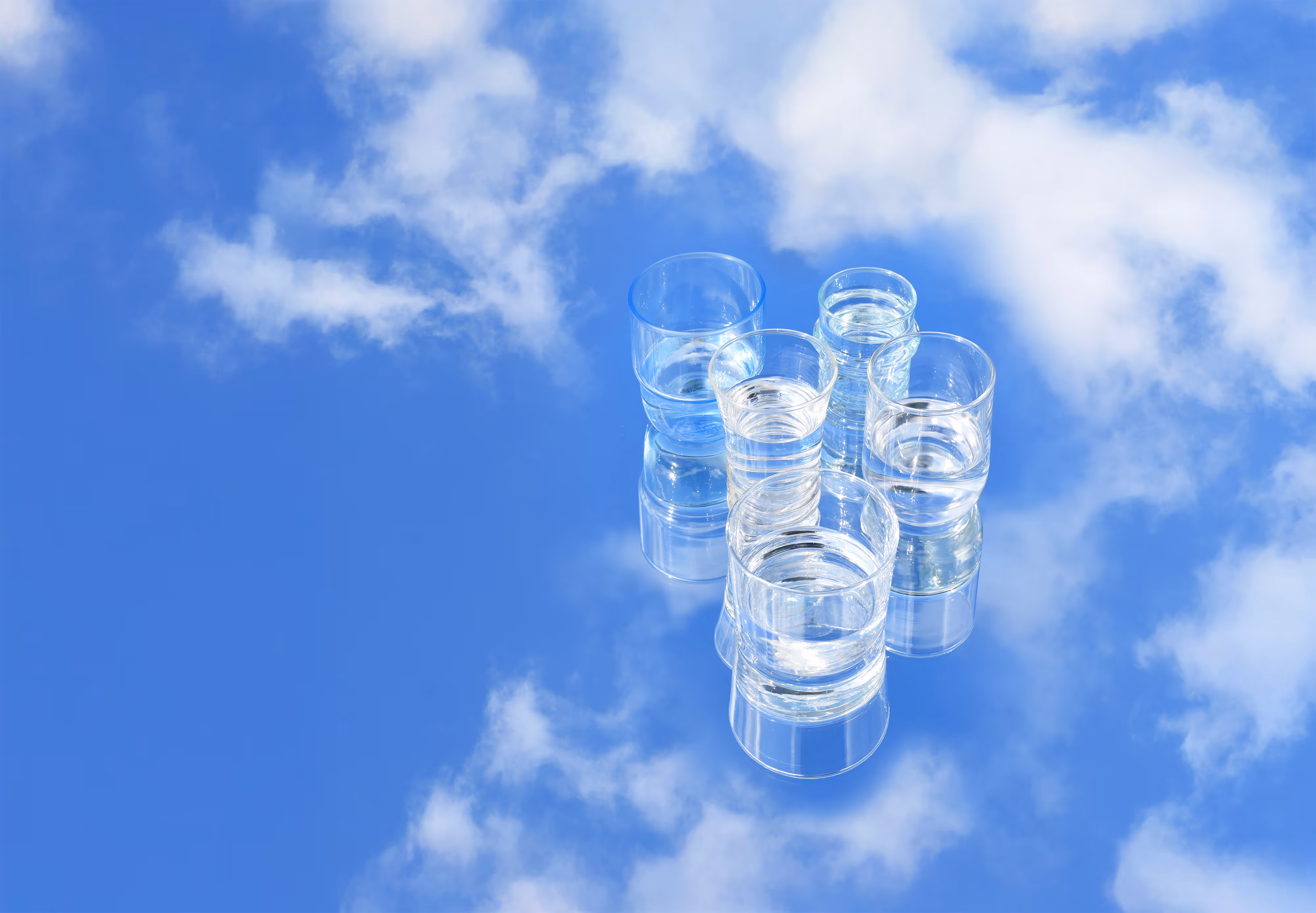 Five glasses of water resting on a mirror on the ground, that's reflecting the blue sunny sky as well as the glasses.