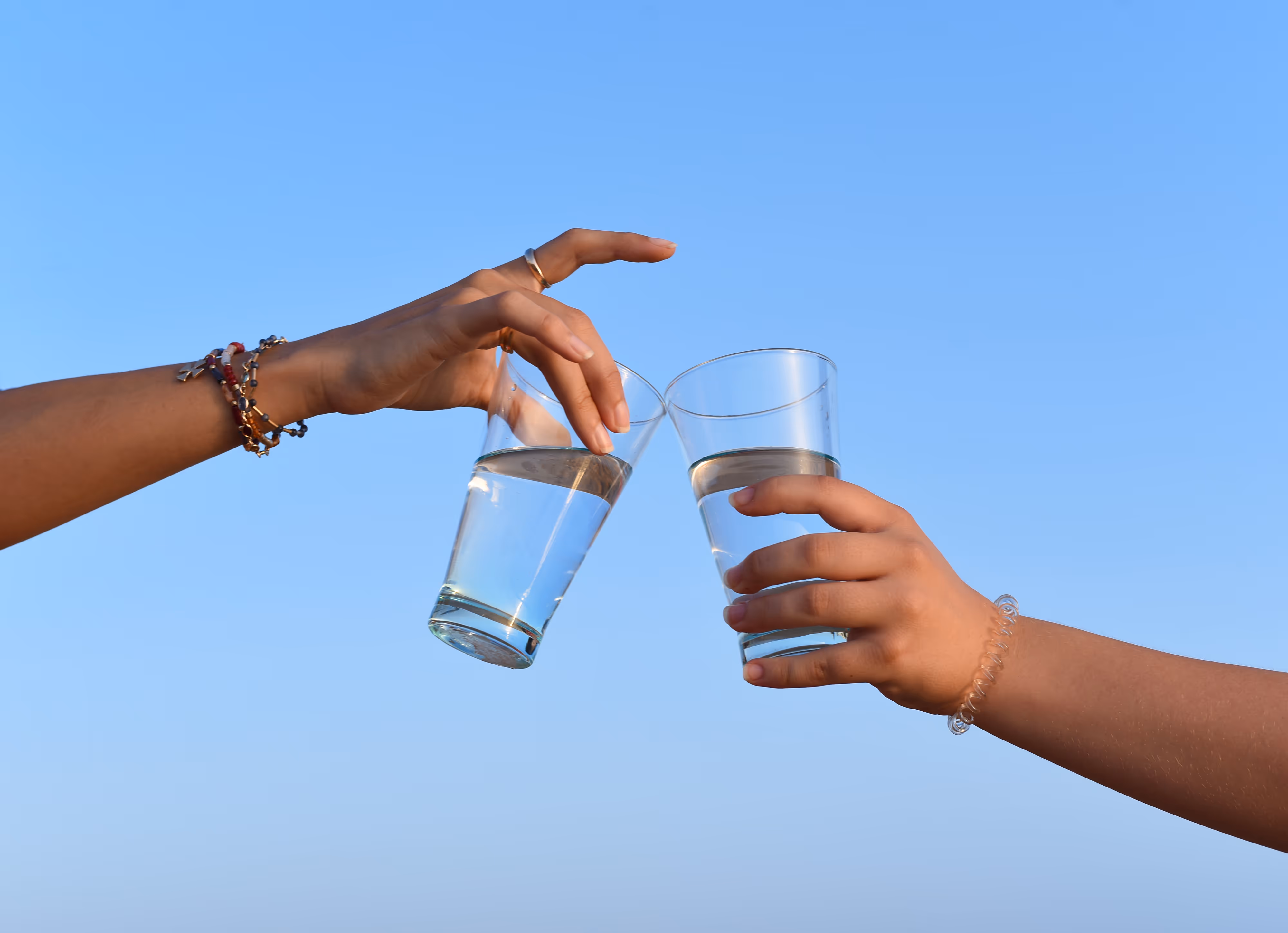 Two hands clinking glasses of water against the backdrop of a clear blue sky.
