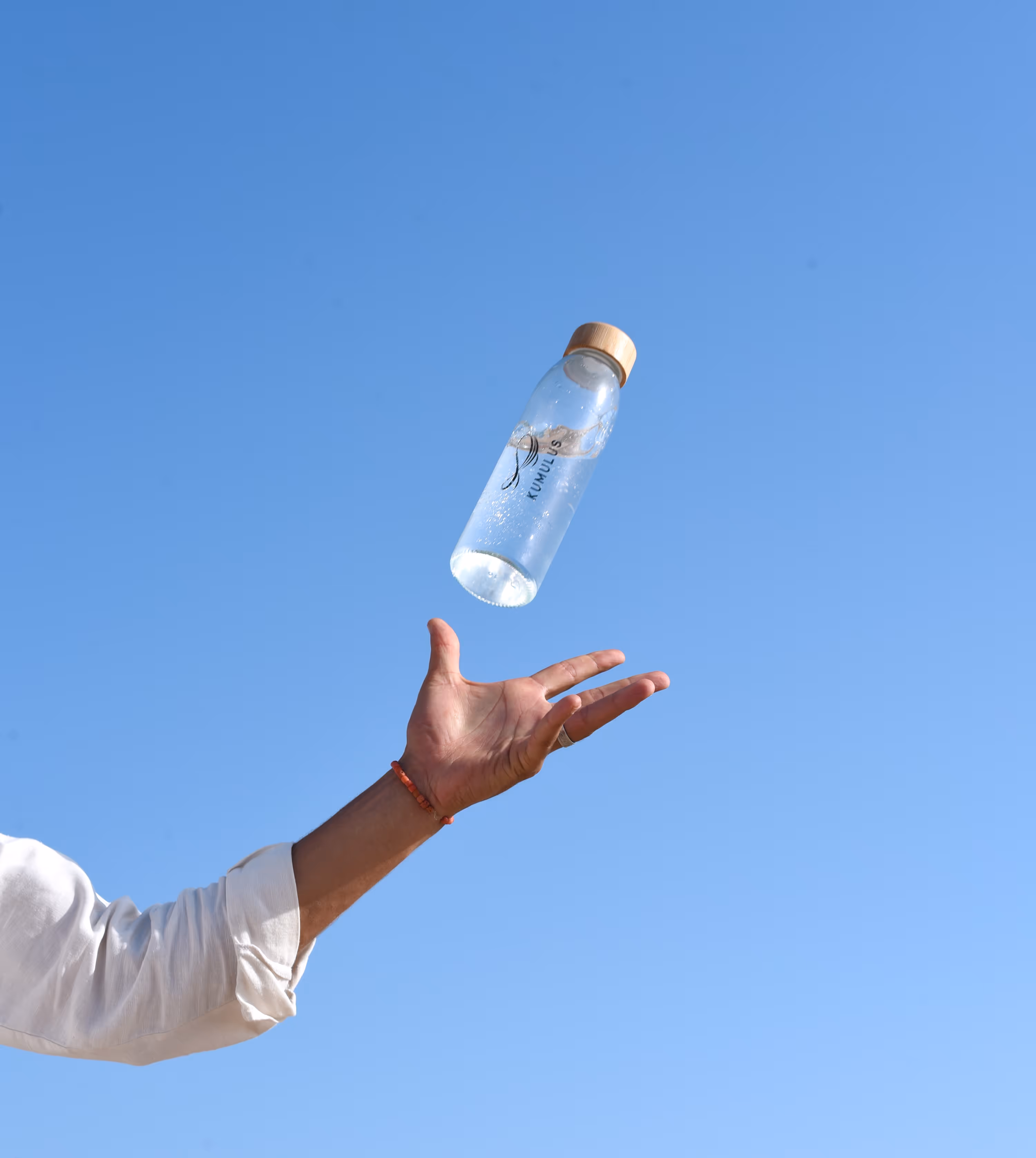 A mans arm tossing a bottle of water, which is mid-air, against the backdrop of a clear blue sky.