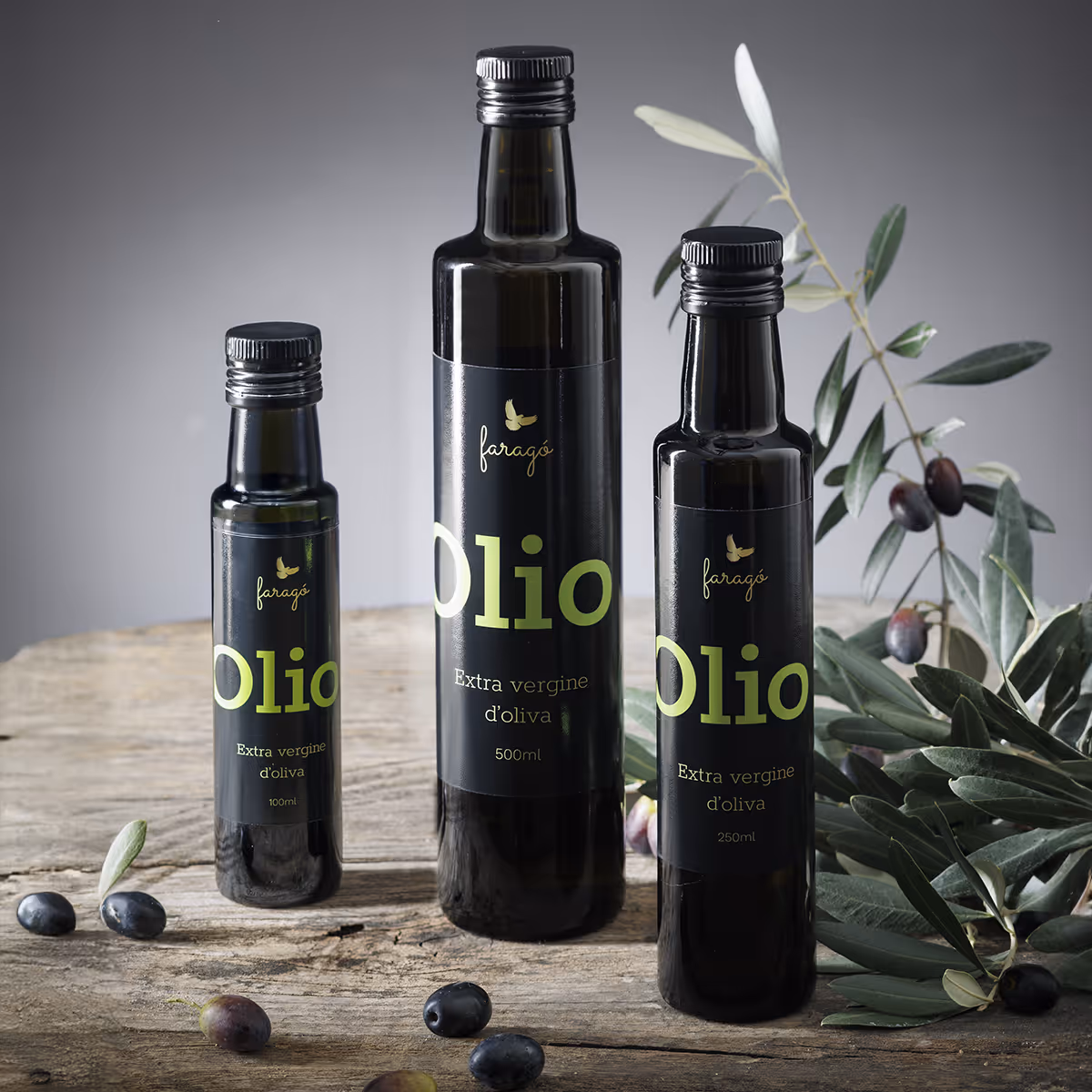 Drei Flaschen natives Olivenöl extra von Faragó auf einer Holzfläche, umgeben von Olivenzweigen und losen Oliven.