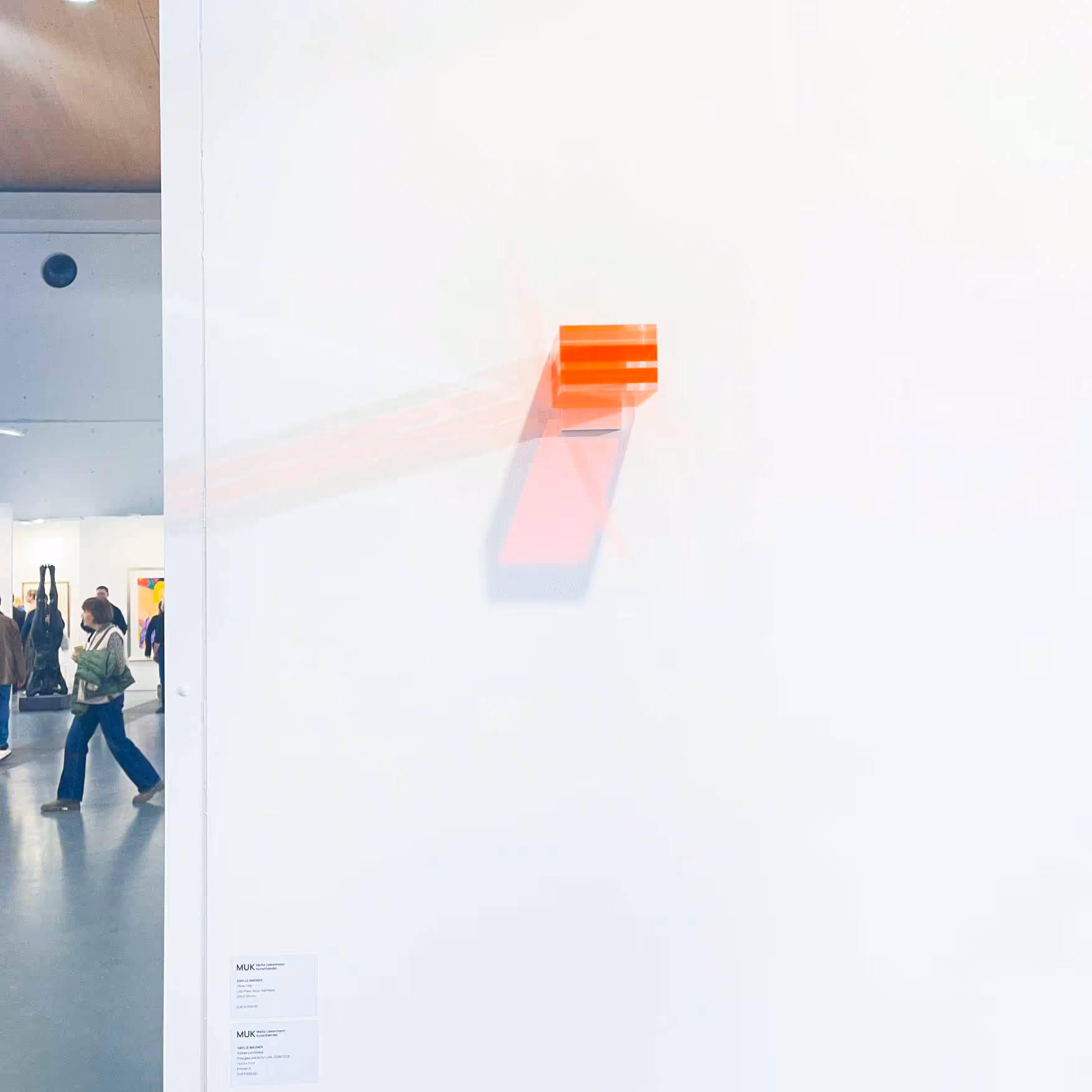 Kleines oranges, transluzentes Kunstobjekt, das an einer weißen Wand in einer Galerie hängt und einen farbigen Schatten wirft.