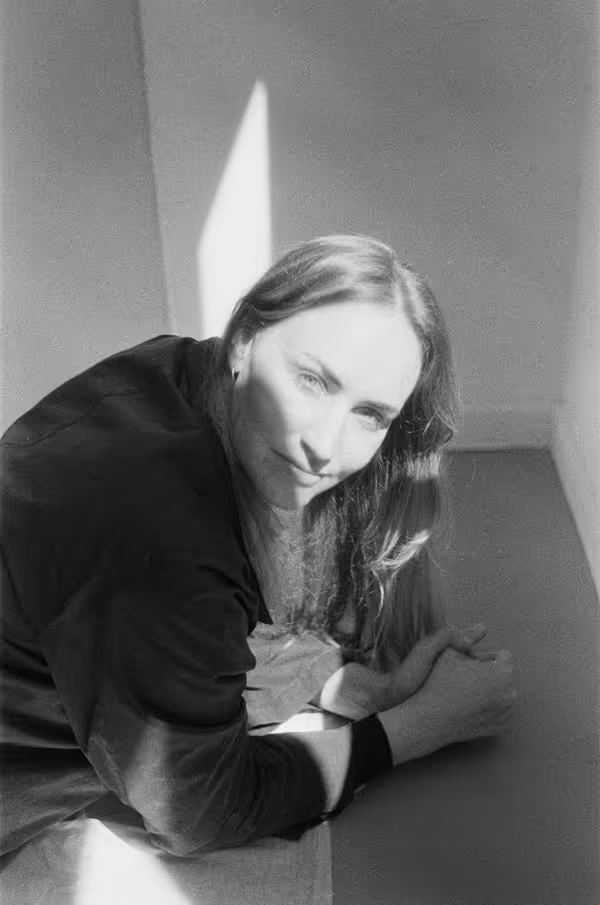Porträt einer Frau mit langen Haaren, die in einem Raum mit Sonnenlicht sitzt und leicht nach vorne geneigt ist von Sophie Rowley