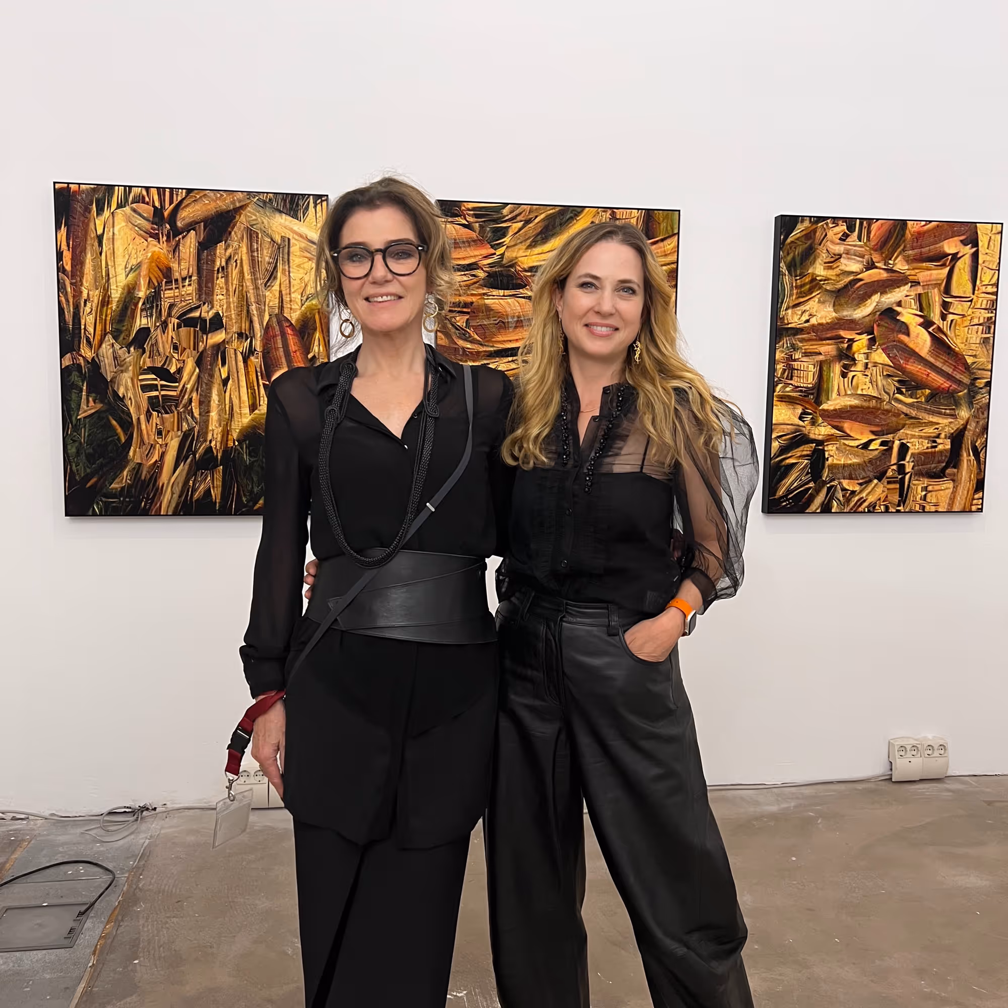 Martina Ziegler und Galeristin Astrid Paul von MUK in schwarzer Kleidung vor abstrakten Gemälden mit goldenen Farbtönen in einer Galerie.