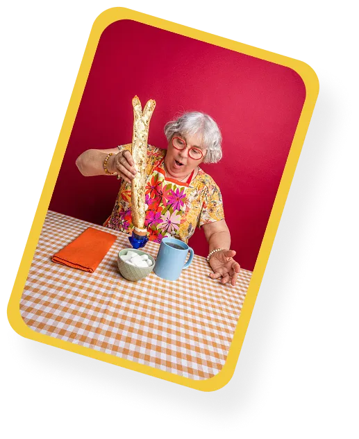 Femme âgée portant des lunettes rouges verse une baguette dans une tasse bleue, assise à une table à nappe à carreaux orange et blanc avec un bol de sucre.