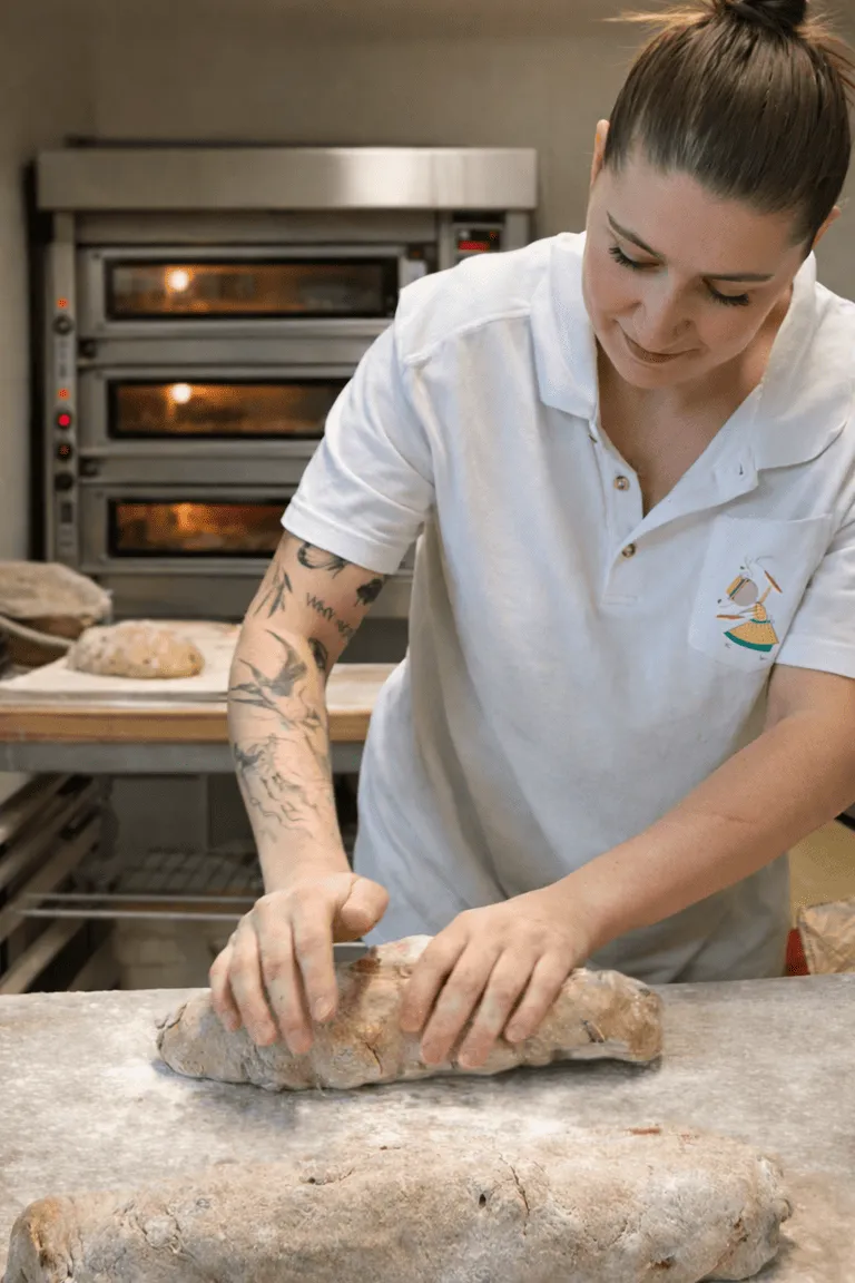 Une boulangère tatouée façonne une pâte à pain sur un plan de travail dans une boulangerie.