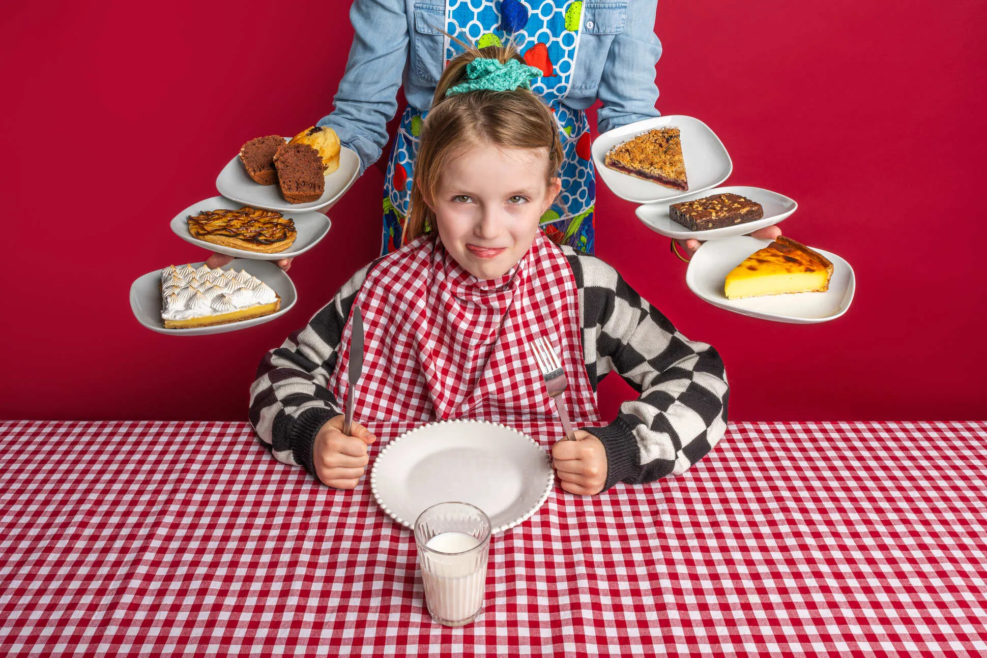 Jeune fille portant un bavoir à carreaux rouges et blancs, tenant un couteau et une fourchette devant une assiette vide, un verre de lait sur une table à nappe à carreaux rouges et blancs, avec une personne derrière tenant six assiettes de desserts variés.