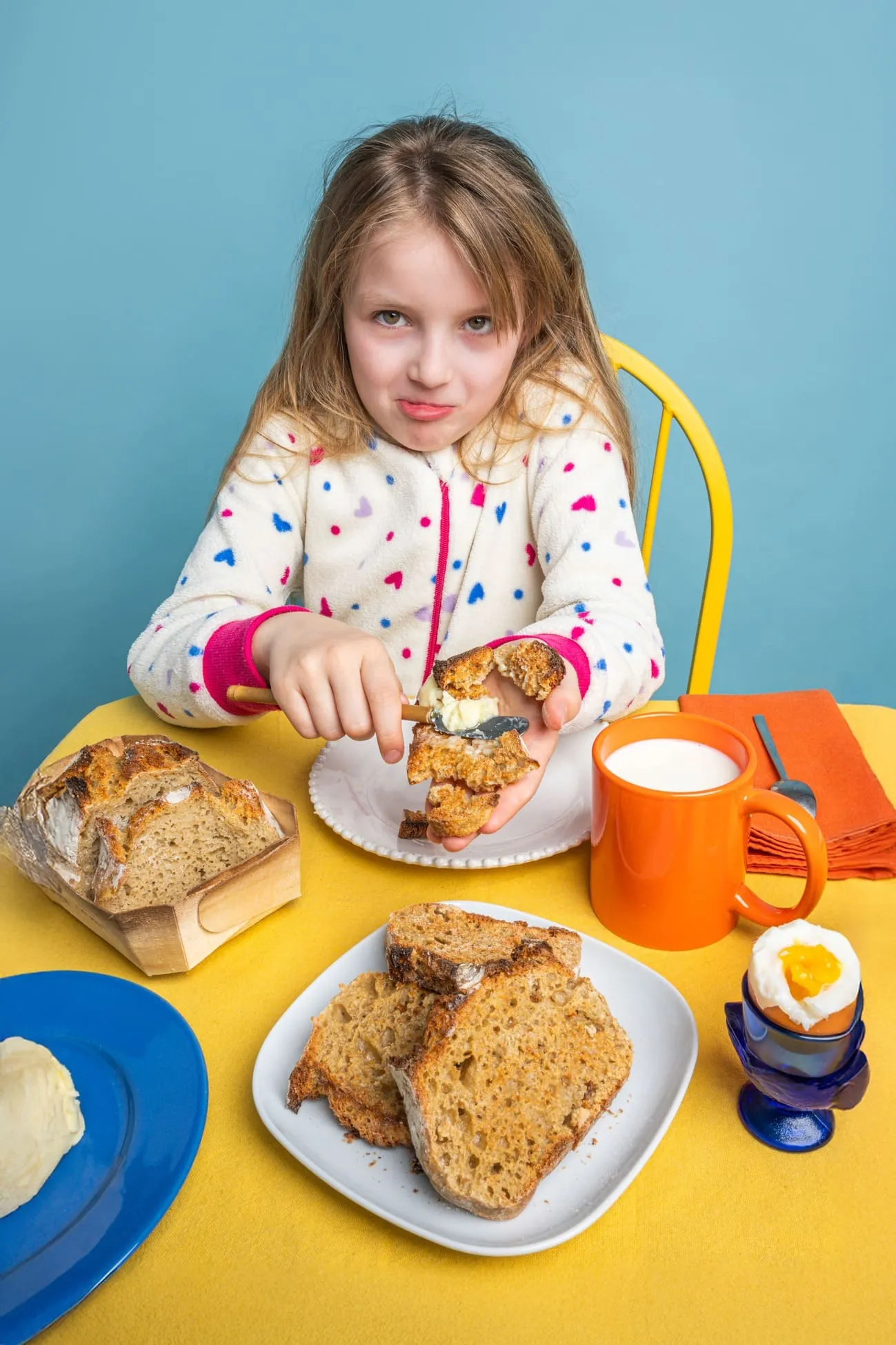 Petite fille tartinant du beurre sur du pain grillé à table avec du pain, un œuf à la coque et une tasse de lait.