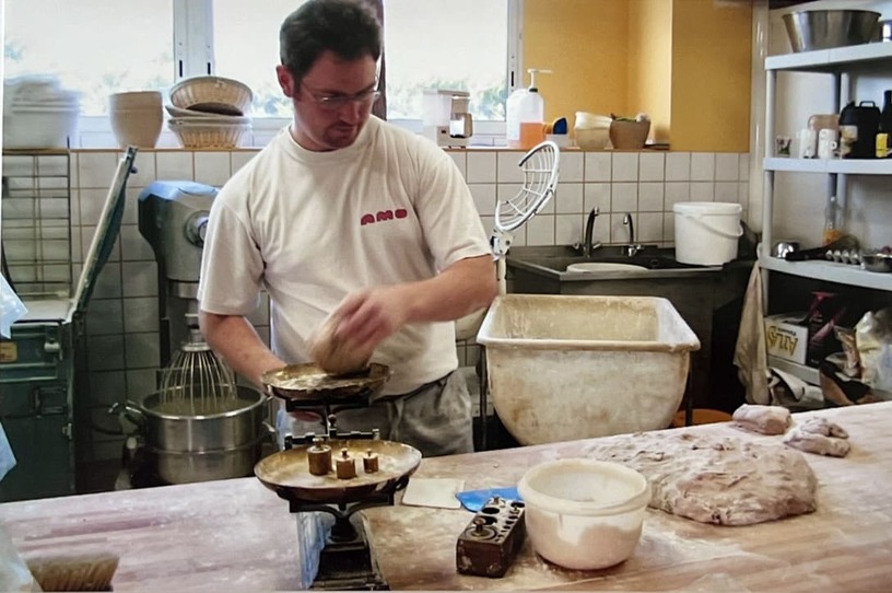 Un boulanger en t-shirt blanc pétrit de la pâte dans un fournil avec des balances anciennes et divers ustensiles de boulangerie autour.