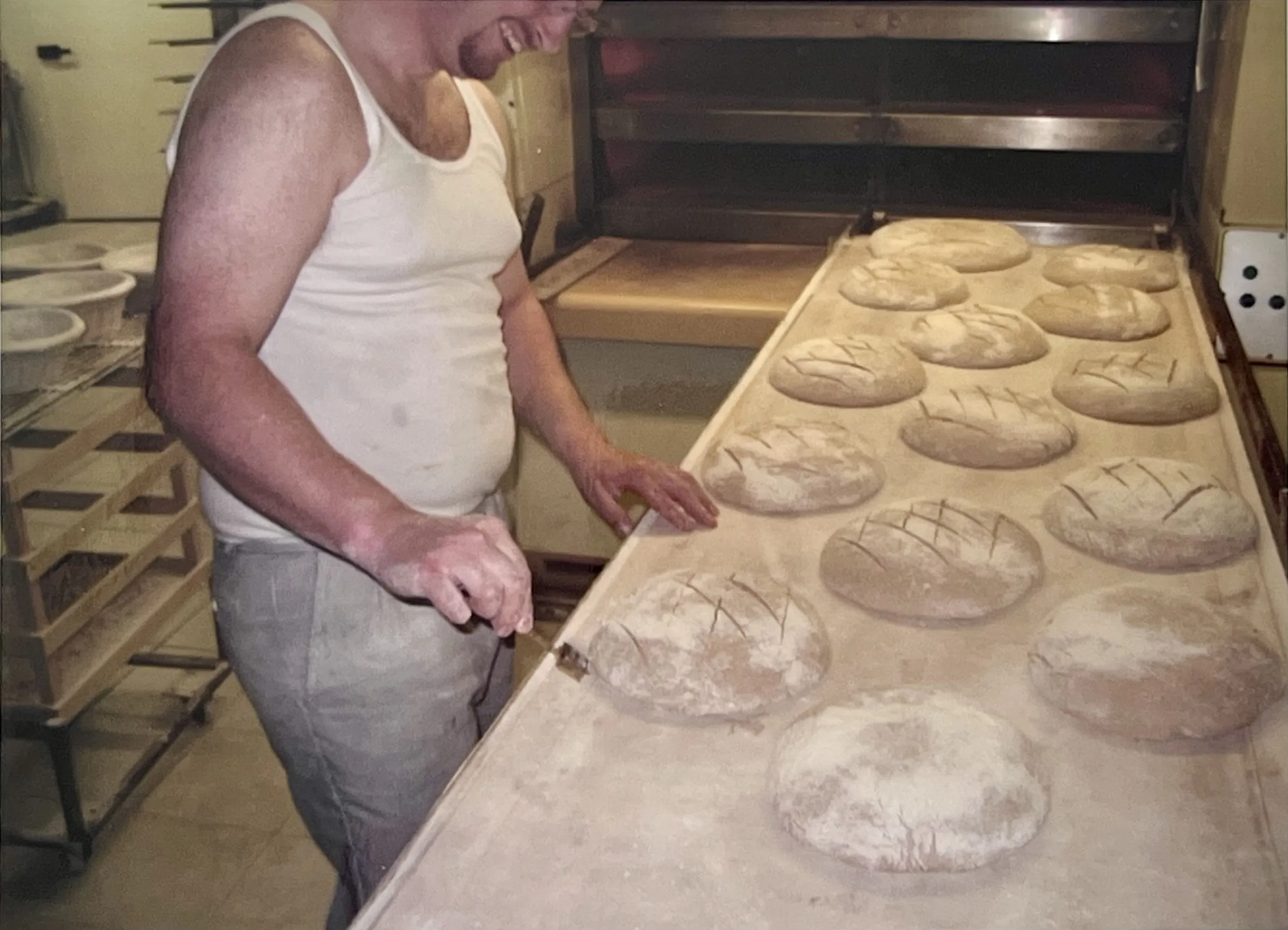 Un boulanger en débardeur blanc souriant prépare des miches de pain avant cuisson dans un four.