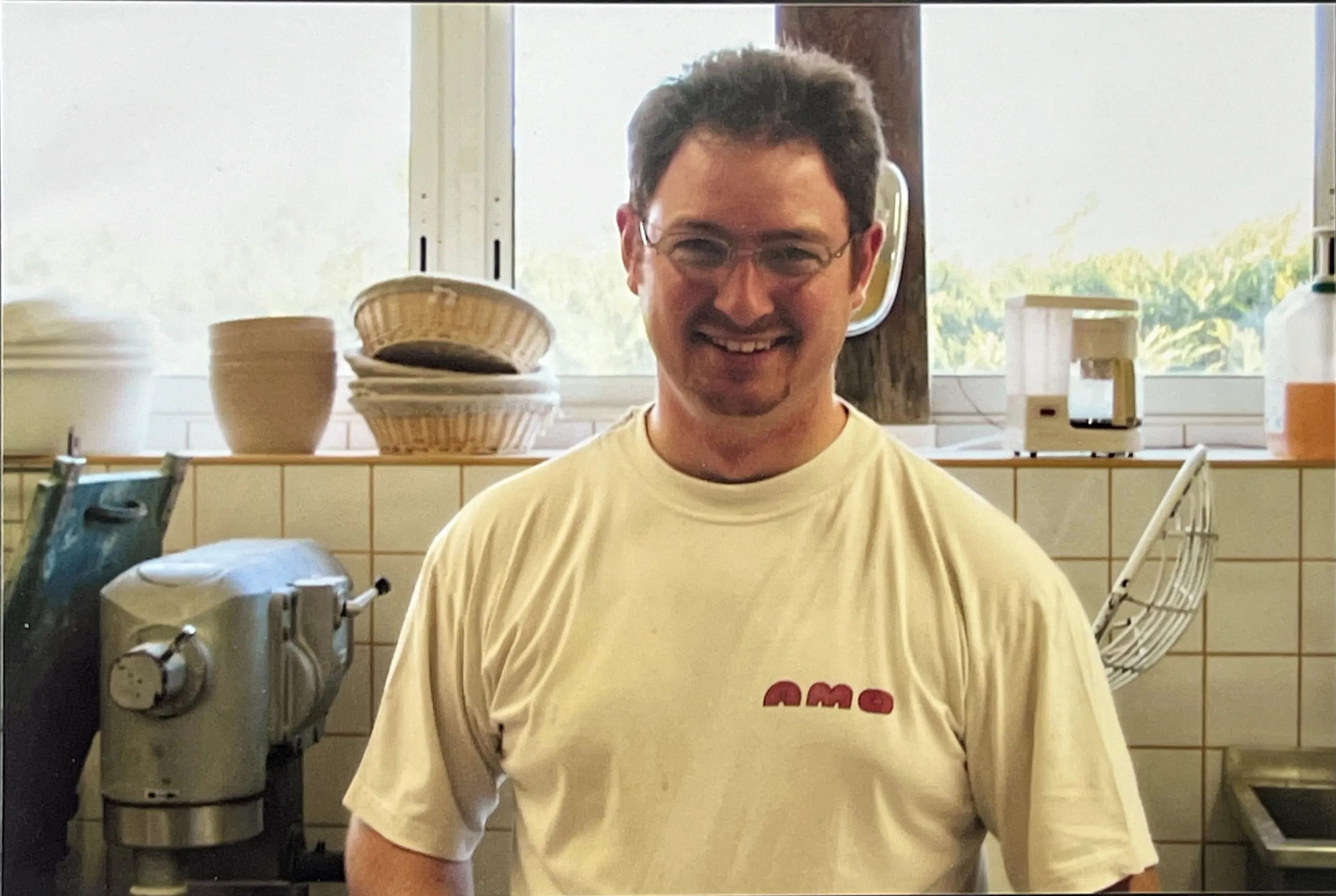 Homme souriant portant un t-shirt blanc avec le logo 'AMO', debout dans une cuisine avec des équipements de boulangerie en arrière-plan.