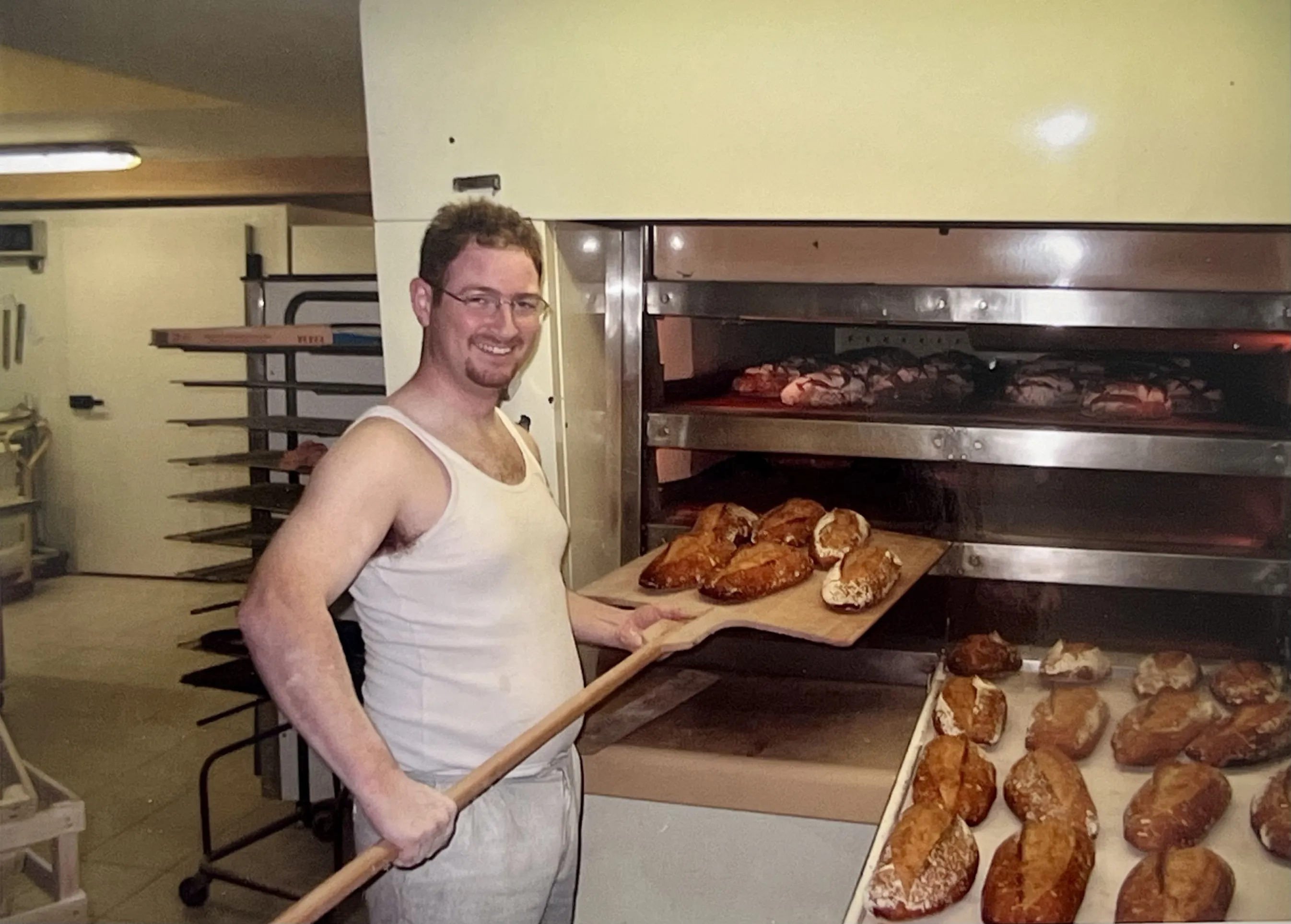 Boulanger souriant en débardeur blanc retirant des pains cuits d'un four industriel dans une boulangerie.