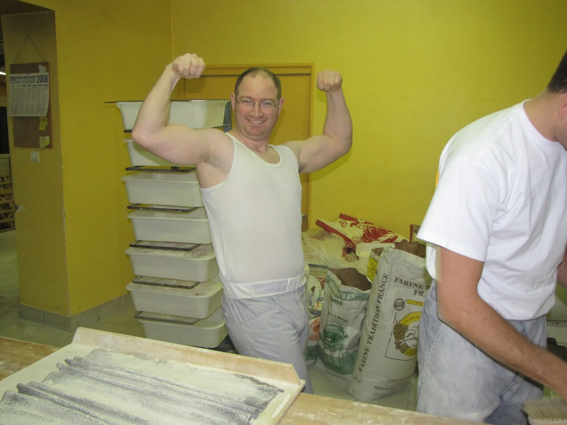 Un boulanger en débardeur blanc souriant et montrant ses muscles dans une boulangerie avec des sacs de farine en arrière-plan.