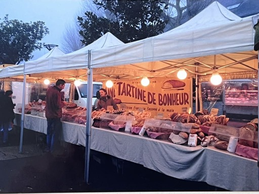 Marché en plein air avec un stand couvert proposant diverses sortes de pain artisanal sous une bannière 'Tartine de Bonheur'.
