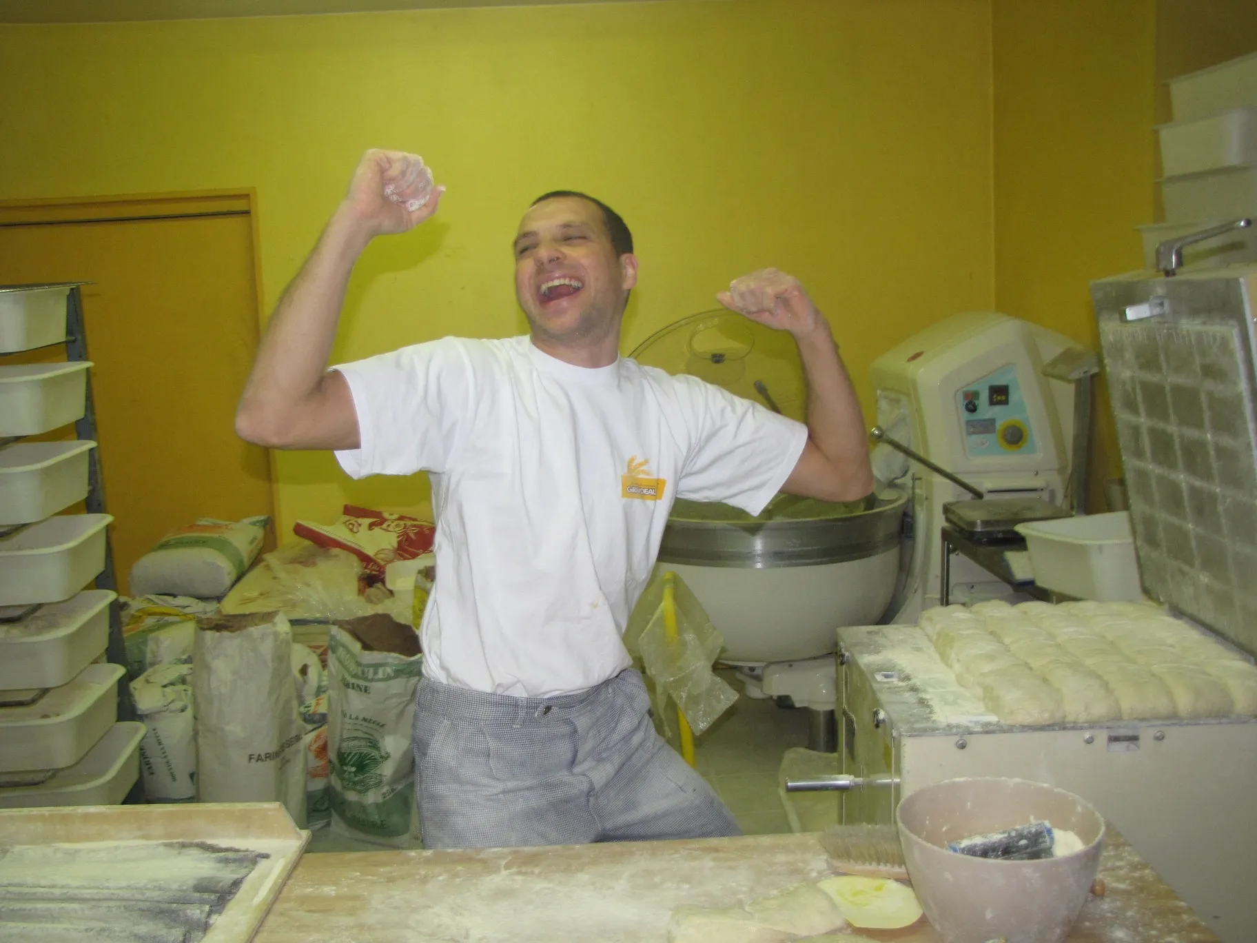 Un boulanger souriant en tablier blanc célèbre dans une boulangerie avec des pâtes à pain sur une table.