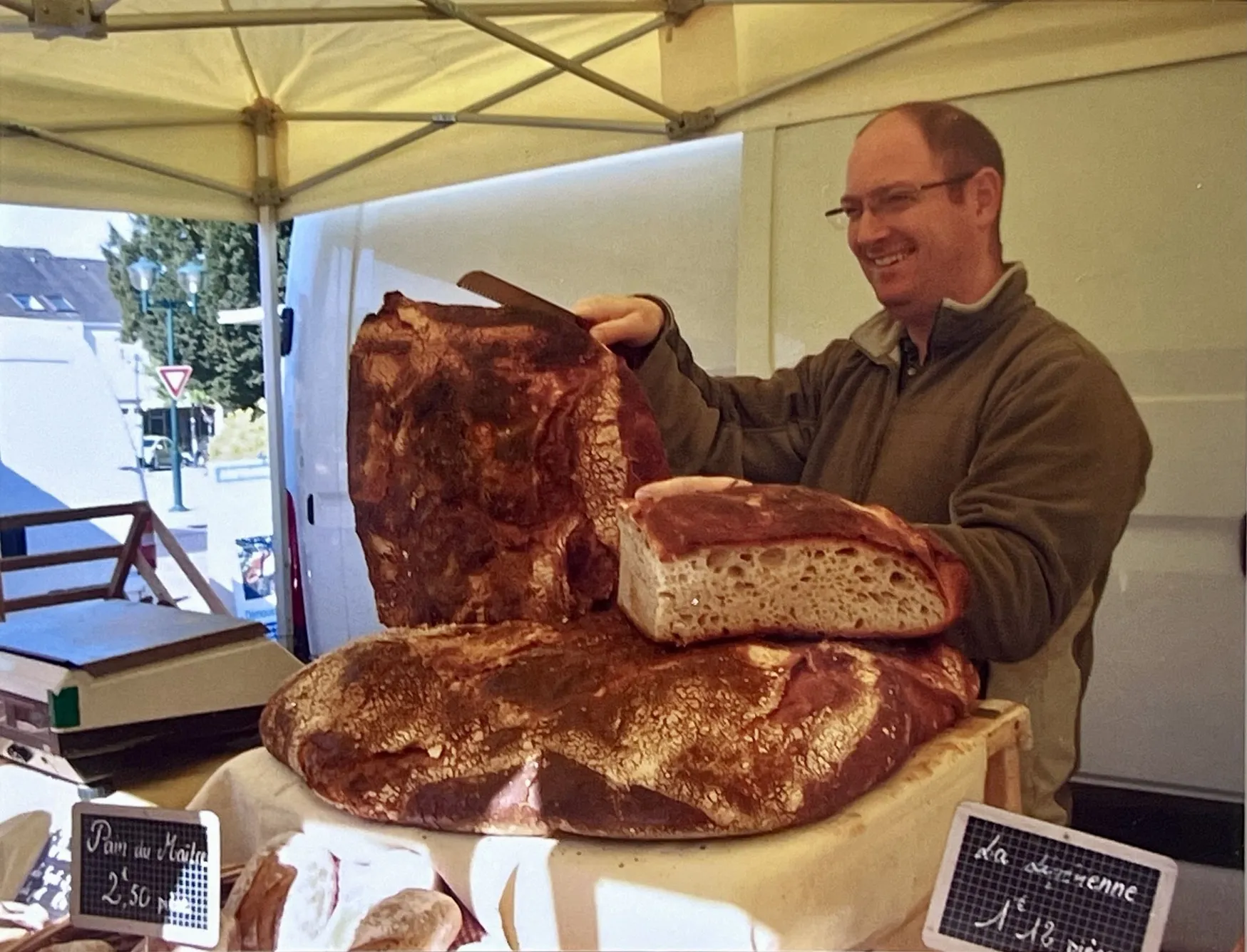 Un homme souriant tenant une grande miche de pain avec une croûte foncée dans un stand de marché sous un auvent.