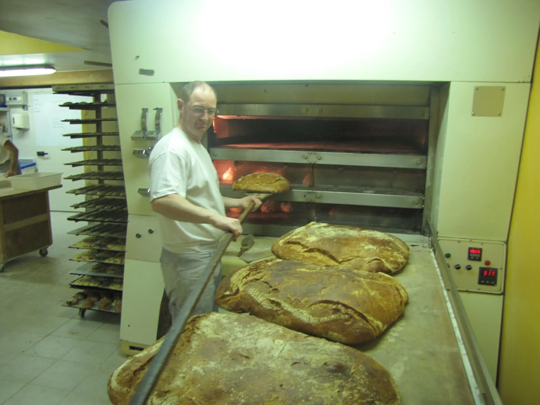 Boulanger utilisant une longue pelle pour sortir un grand pain du four industriel dans une boulangerie.