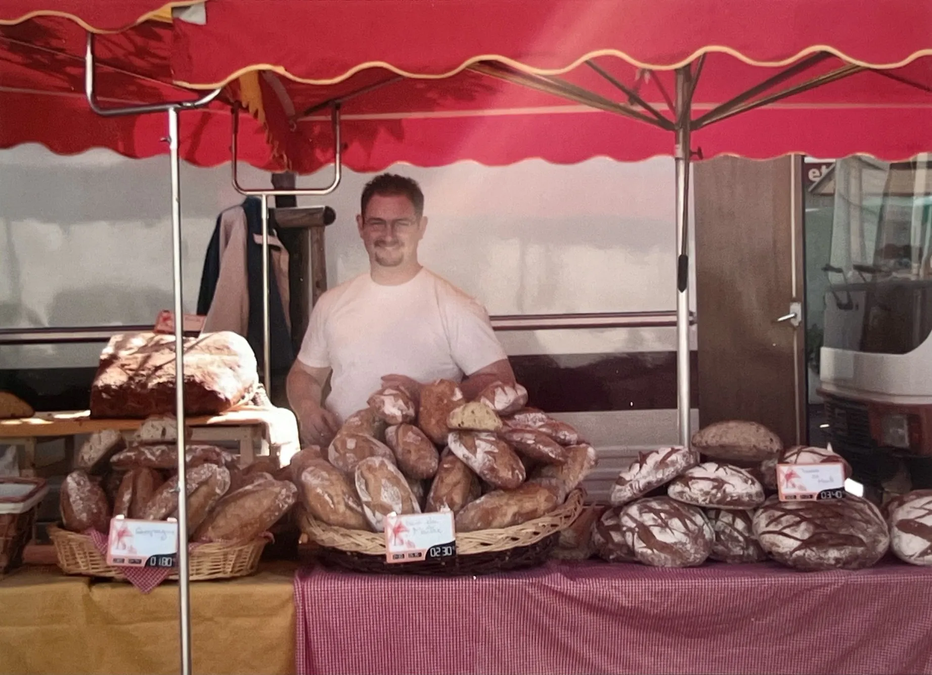 Homme souriant en t-shirt blanc debout derrière un stand avec une variété de pains sur une table sous un parasol rouge.