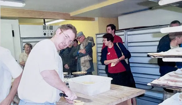 Un homme en t-shirt blanc tranche de la pâte sur une table dans une boulangerie avec plusieurs personnes en arrière-plan.