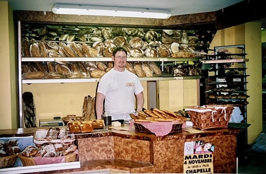 Homme souriant dans une boulangerie avec des étagères pleines de pains et une caisse en bois devant lui.