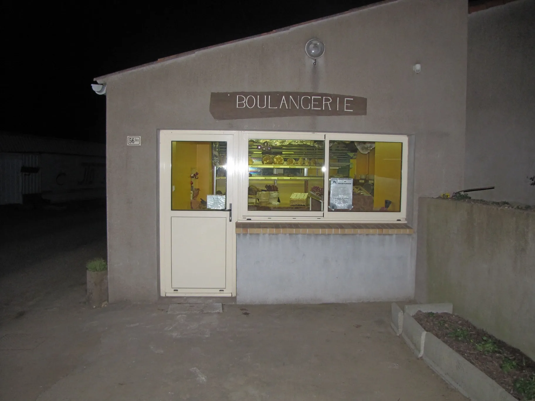 Façade d'une petite boulangerie éclairée de nuit avec porte blanche et vitrine montrant des pains à l'intérieur.