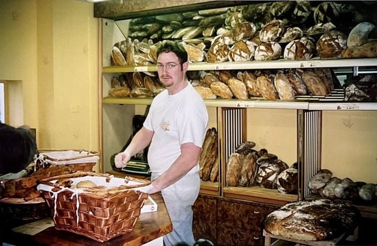 Un boulanger portant un t-shirt blanc travaille dans une boulangerie avec des étagères remplies de pains artisanaux en arrière-plan.