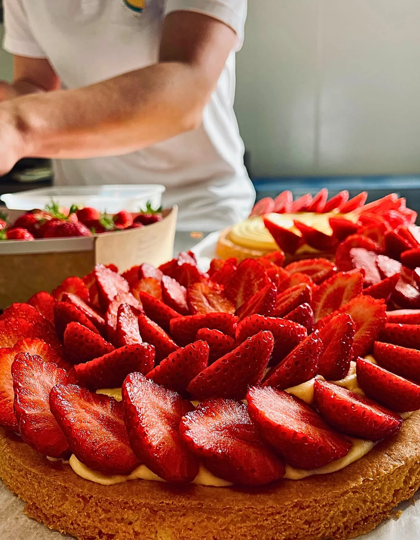 Tarte avec une généreuse couche de fraises tranchées sur une base biscuitée, en arrière-plan une personne en blanc.