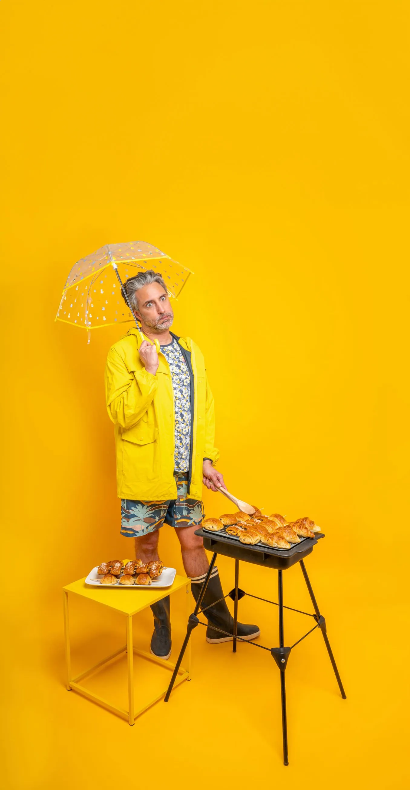 Homme en imperméable jaune et bottes tenant un parapluie transparent près d'un grille-pain avec des croissants.