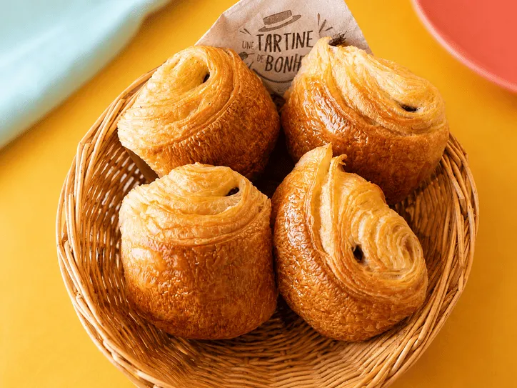 Quatre pains au chocolat dorés dans un panier en osier sur une table jaune.