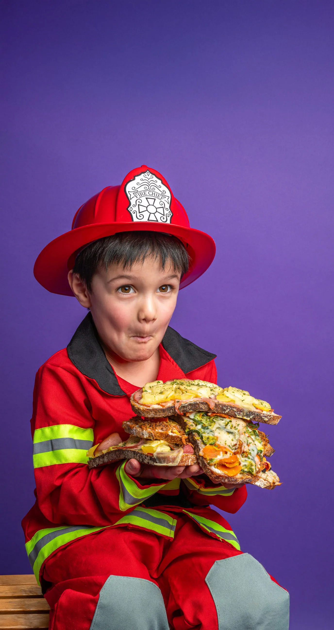 Enfant déguisé en pompier tenant plusieurs tartines garnies avec expression malicieuse.