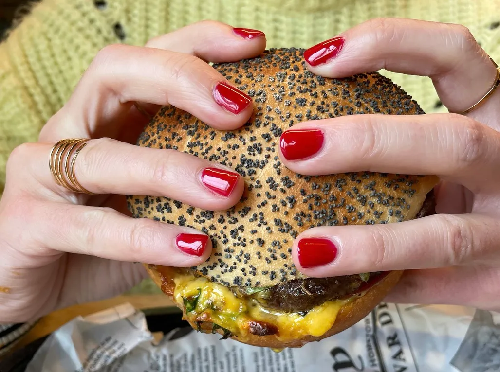 Deux mains aux ongles rouges tiennent un burger au pain saupoudré de graines de pavot.