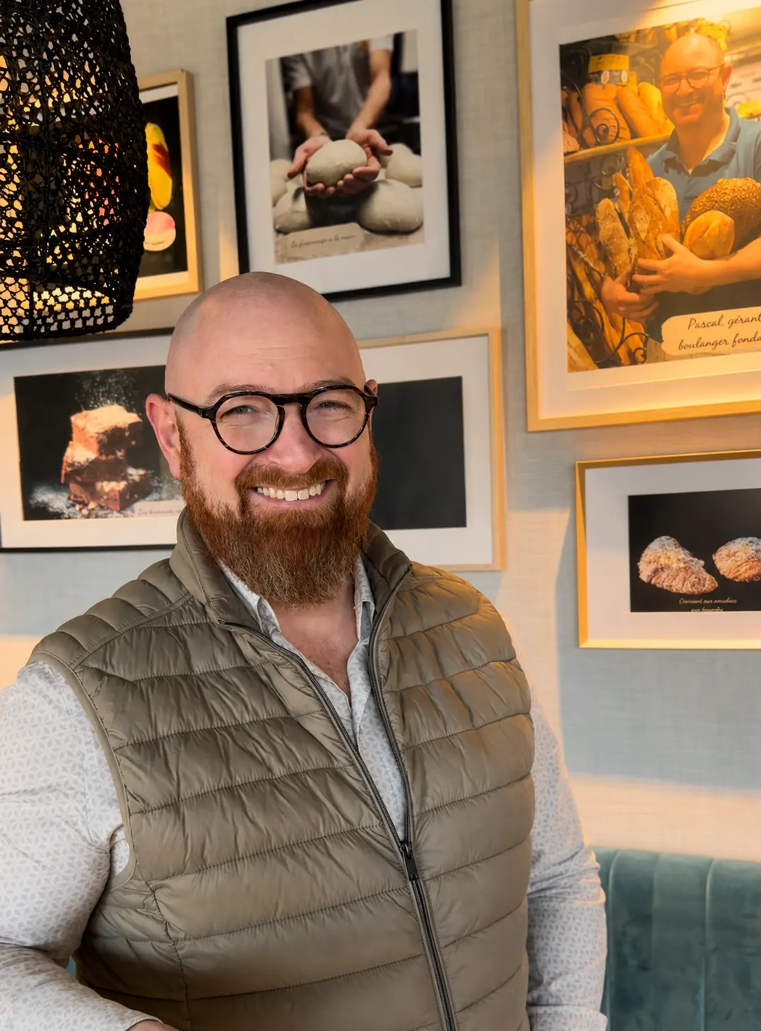 Homme chauve souriant avec une barbe et des lunettes devant un mur avec des cadres de photos de pain.